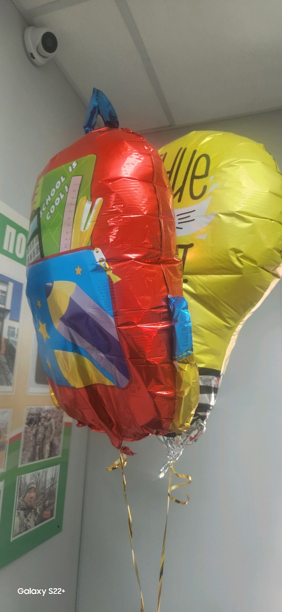 The image shows two helium balloons hanging indoors. The balloon in the foreground is red and decorated with school-themed imagery, including a ruler, pencils, and the text "SCHOOL IS COOL!". The second balloon is yellow and appears to be shaped like a lightbulb with some text that is difficult to decipher. The background is a light-colored wall, and to the left, there are framed pictures on a wall, possibly in a school or office environment. A security camera is visible on the ceiling in the upper left corner. The bottom left corner of the image displays "Galaxy S22+". The scene is static and does not depict any people or actions. The location is stated to be Nyurba, Russia, but this information is not visually verifiable from the image itself.