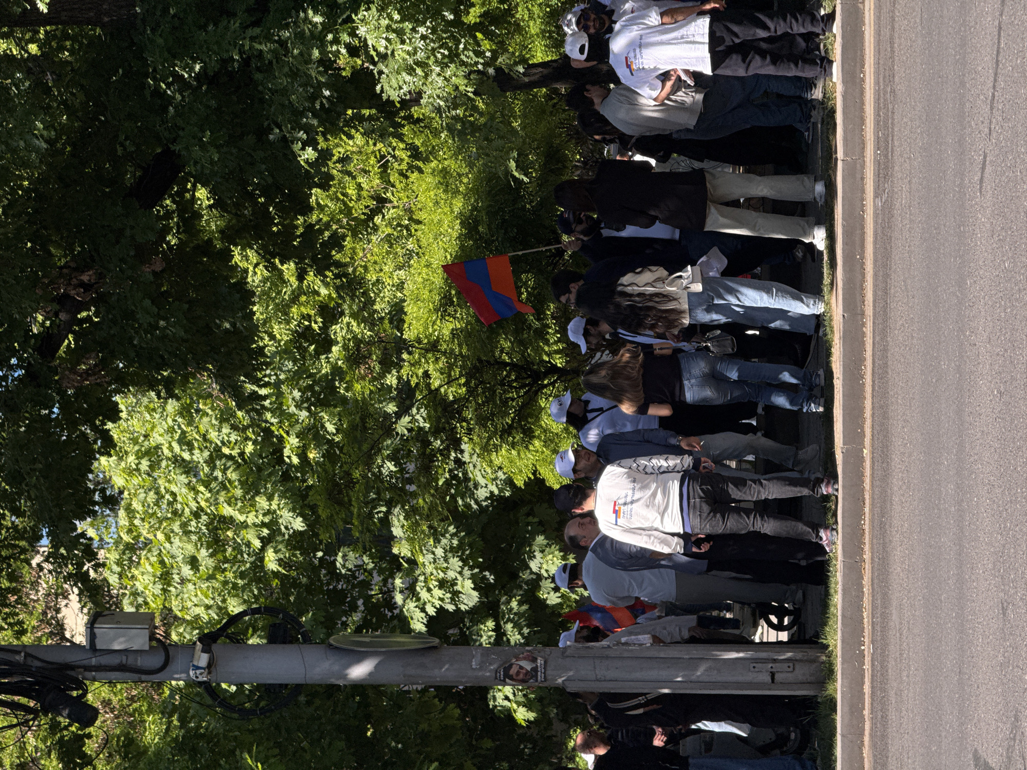 A group of people are gathered on the side of a street in Yerevan, Armenia. They are standing near a line of trees and a sidewalk, with the roadway to their right. Some people in the group are wearing white shirts with graphics, and one person is holding an Armenian flag. A traffic light and utility pole are visible in the foreground.
