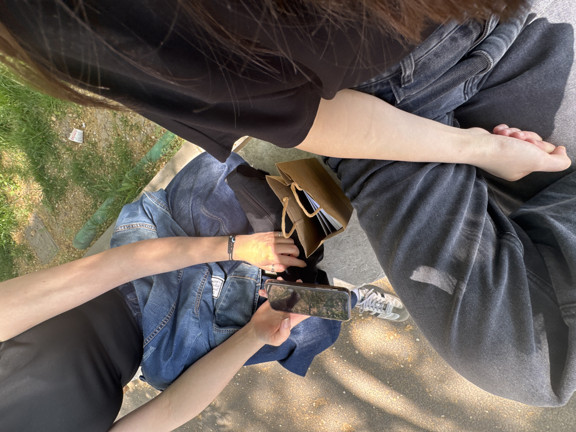 Two people are seated outside, on what appears to be a concrete surface. One person is wearing dark-colored jeans and a black shirt; their legs are crossed at the knees, with the right leg on top. The other person is also wearing a black shirt, with a blue denim jacket spread over their lap. They are holding a mobile phone in their hands. A brown bag sits between the two people. The ground is partly grass. The location cannot be identified with certainty.
