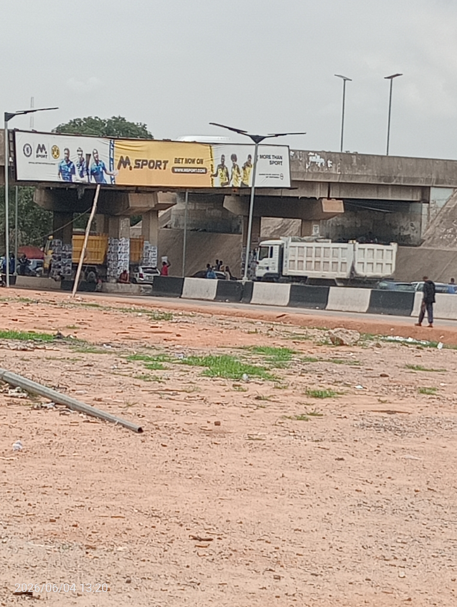 The image captures an outdoor scene beneath a concrete overpass, suggestive of a commercial or transit area, likely in Madalla, Nigeria. A road runs under the overpass, flanked by dirt and gravel foreground.
Two large billboards are affixed to the underside of the overpass. The left billboard displays the "M SPORT" logo alongside a "BVB SPORT" emblem, featuring male athletes in sports jerseys. Prominent text reads "BET NOW ON WWW.MSPORT.COM". The right billboard also shows an "M SPORT" logo and the slogan "MORE THAN SPORT," with more images of athletes.
On the left side of the road beneath the overpass, a yellow dump truck is parked, loaded with numerous packaged goods, which appear to be white jerry cans. Several people are visible around this truck, some sitting, others standing. A white dump truck is also seen on the right side of the road, driving away from the camera. Further right, a person in dark attire walks along the roadside.
The foreground consists of reddish-brown, uneven dirt and gravel, with sporadic patches of green vegetation. A metal pole or pipe lies horizontally in the lower left. The sky is uniformly light and overcast. A timestamp in the lower left corner indicates "2026/06/04 13:20," suggesting the photograph was taken in the early afternoon. The scene conveys a sense of bustling activity in a functional, semi-urban setting.