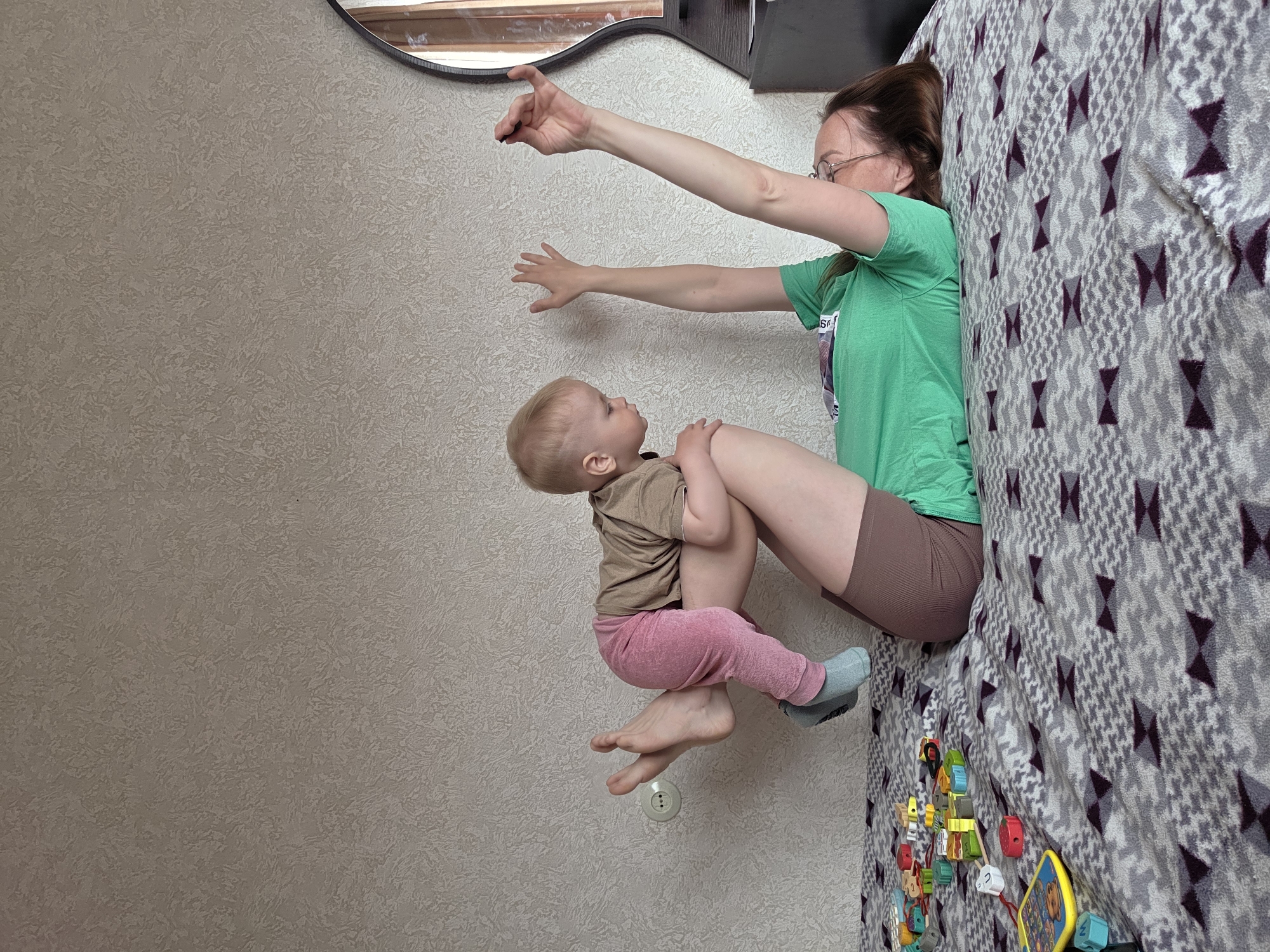 A woman with long brown hair and glasses is seated on a bed covered with a grey and white patterned blanket. She is wearing a green t-shirt and brown shorts. She is holding a baby, dressed in a brown shirt and pink pants, in her arms. Her arms are outstretched. The scene takes place indoors, next to a wall with a neutral textured surface. Scattered on the bed near the woman are colorful children's toys. A partial view of a dark-framed mirror is visible in the upper part of the picture. The location of the image is the city of Chaikovsky, Russia.