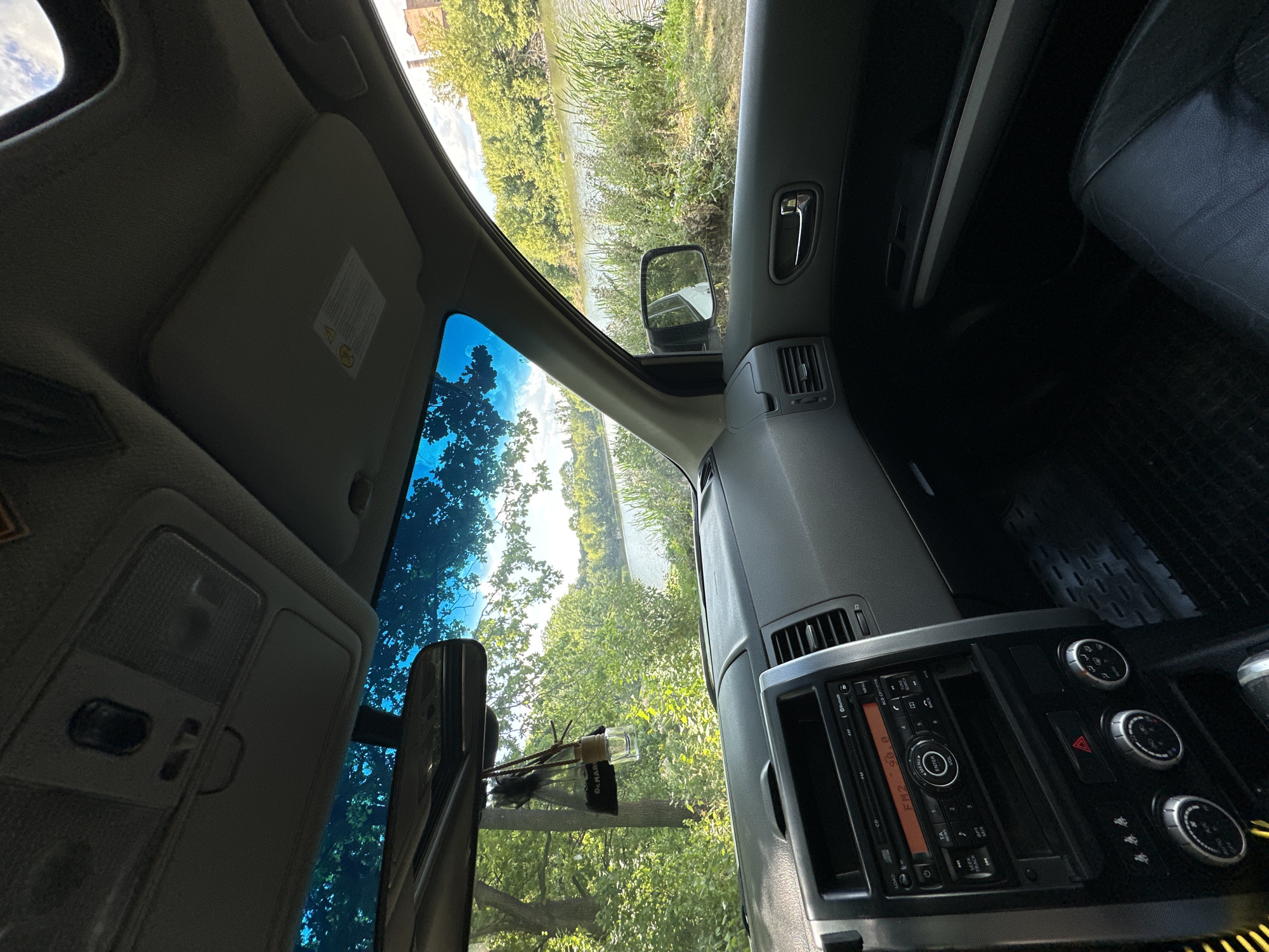 The image shows the interior of a car, looking out towards a body of water surrounded by trees. The dashboard of the car is visible in the foreground, with the radio and climate controls. A car air freshener hangs from the rearview mirror. Through the windshield, a lake or river can be seen, with lush green trees lining its banks. The sky is bright and partly cloudy. The overall scene suggests a peaceful, natural setting. It is not possible to determine the location in Shatava, Ukraine from the image.
