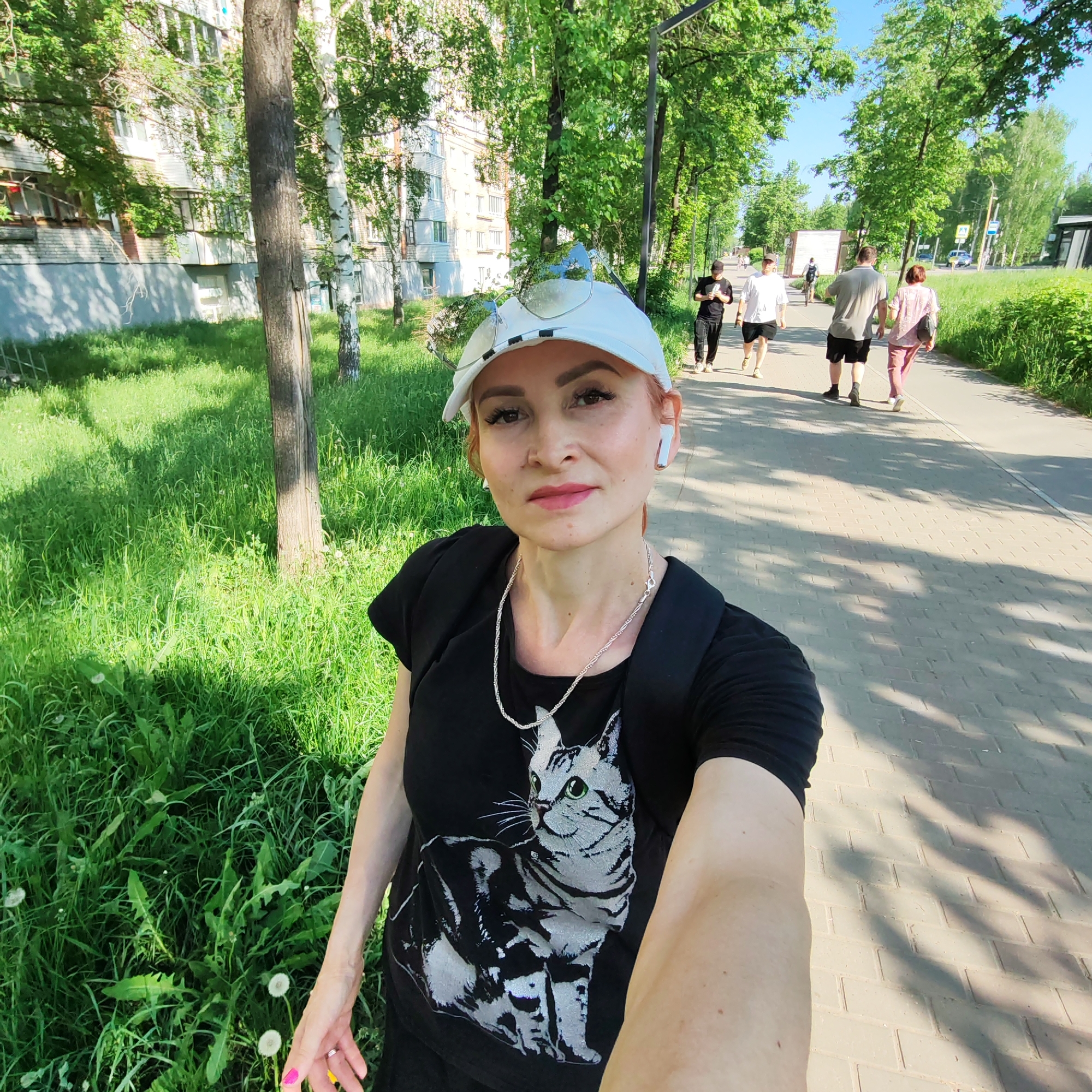 A fair-skinned woman with light red hair, wearing a white baseball cap with sunglasses perched on top, a black t-shirt with a cat graphic, and a silver necklace, is taking a selfie on a paved walkway. She is wearing wireless earbuds in her left ear. The walkway is lined with green grass and trees on the left and more trees and green space on the right. In the background, several people are walking along the walkway in the same direction, away from the camera. In the far background, there is a multi-story building on the left and a smaller building on the right. The scene appears to be outdoors during daylight hours. The location is Chaikovsky, Russia.