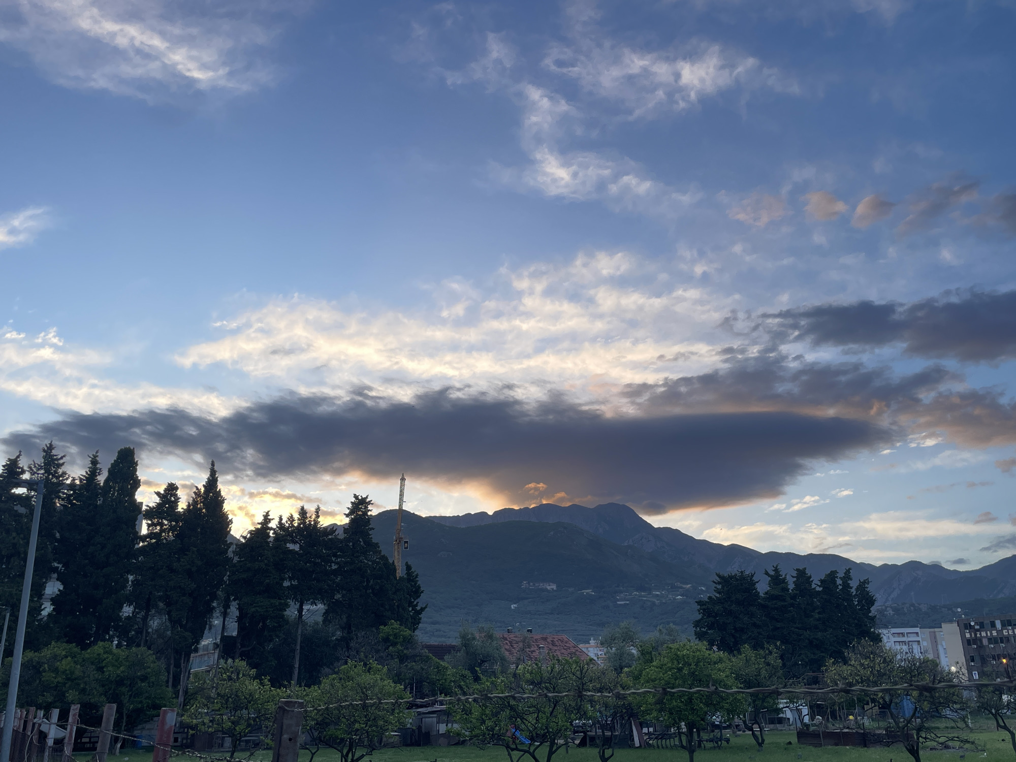 The image depicts a landscape in Bar, Montenegro, featuring a mountain range in the background partially obscured by a long, dark cloud formation tinged with orange light. In the foreground is a fenced green area with a variety of trees and vegetation, including fruit trees. Behind the fence, there is a structure with a red tiled roof, partially hidden by trees. A construction crane is visible among the trees. To the right, several buildings can be seen. The sky is a mix of blue and white, with scattered clouds.