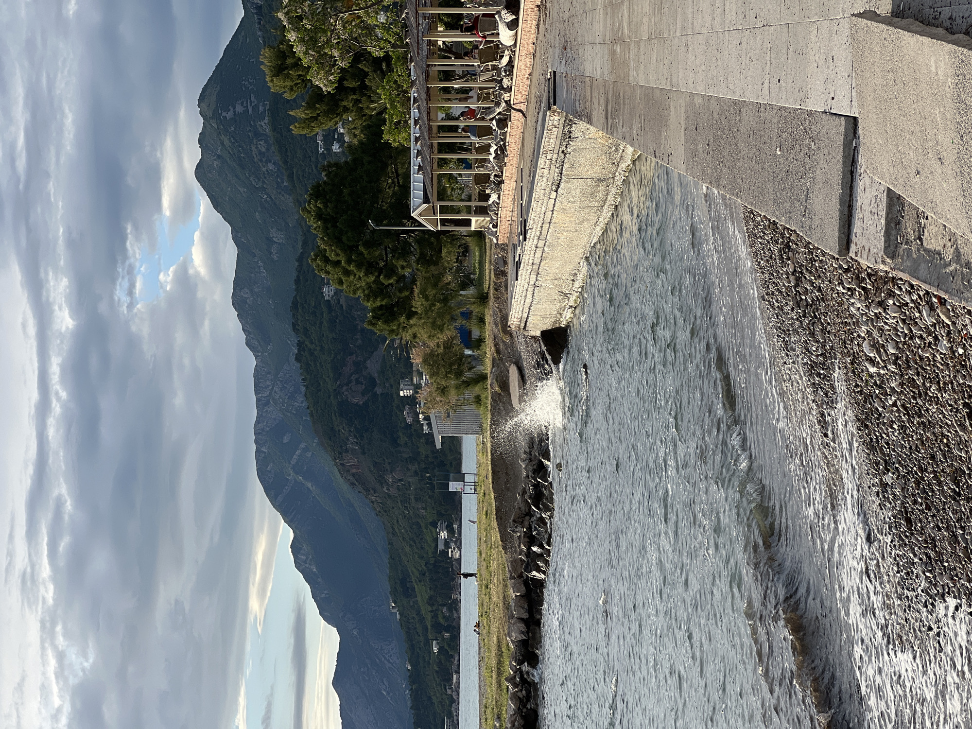 The image captures a seaside scene in Bar, Montenegro. A mountain rises prominently in the background under a cloudy sky. Adjacent to the water is a concrete embankment and a rocky shoreline. Water is crashing against the shore. Near the embankment, there's a restaurant or outdoor seating area with chairs and tables. Trees grow nearby. In the distance, there appear to be people standing along the shoreline of the bay.