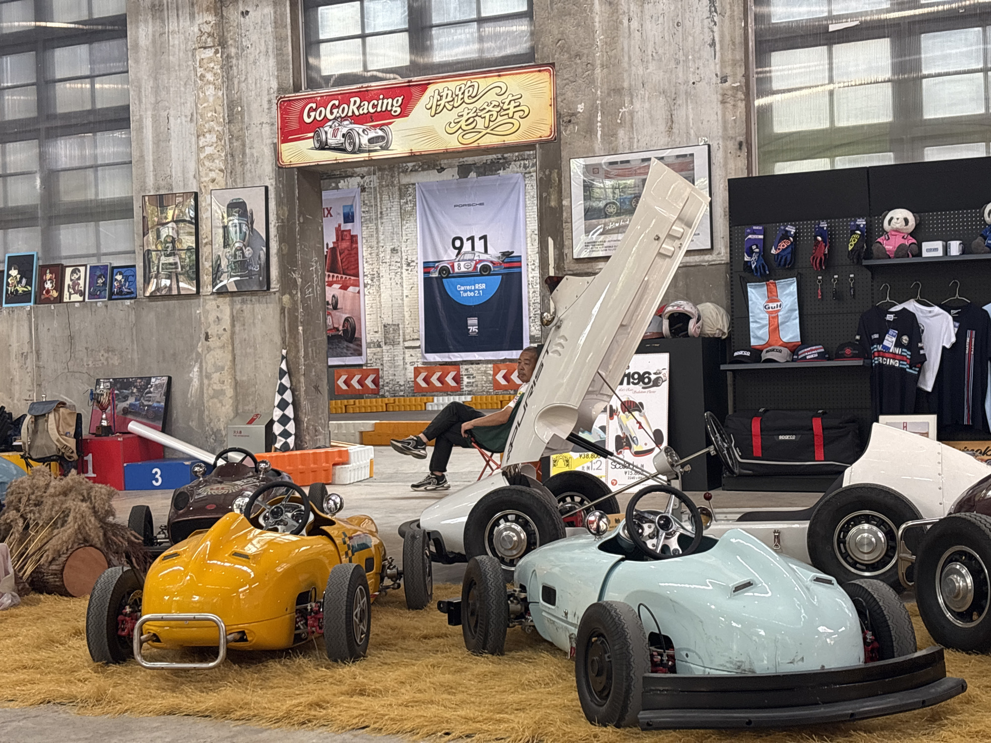 The image depicts a display of vintage-style go-karts and automotive memorabilia, likely in a retail or exhibition space within Pudong, China. In the foreground, three go-karts are arranged on a textured yellow mat: a bright yellow one on the left, a light blue one in the center, and a white one partially visible on the right, with a dark brown one further back. A man is seated on a red folding chair in the middle ground, leaning against the open hood of the white go-kart. Behind the go-karts, shelves are stocked with merchandise including gloves, hats, and t-shirts, featuring brands like Gulf and Sparco. Automotive-themed posters and banners adorn the concrete wall in the background, including a large Porsche 911 banner and a vintage "GoGoRacing" sign. The environment appears to be an indoor space with a concrete wall and large windows with visible mullions.