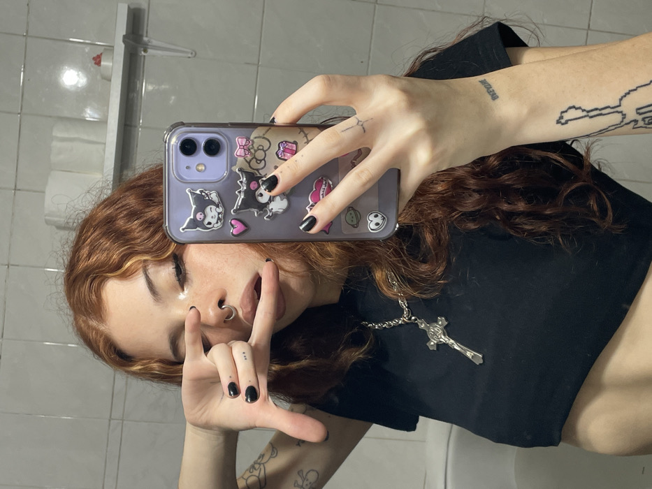 A young individual with long, wavy reddish-brown hair featuring blonde streaks is depicted taking a mirror selfie. The subject wears a black crop top and a silver-colored chain necklace with a crucifix pendant. Facial features include visible freckles, winged eyeliner, a septum piercing, and a bright highlight on the nose bridge. The subject's left hand is raised, with the index finger and thumb forming a rectangular gesture, partially obscuring the left eye, while the mouth is slightly open with the tongue visible. The right hand holds a mobile phone, an iPhone model, encased in a clear purple case adorned with multiple cartoon stickers, predominantly depicting Kuromi characters, a pink heart, and a pink bow. Both hands display black nail polish. Tattoos are visible on the left forearm (pixelated guitar outline), left wrist (small text), left ring finger (three dots), and the back of the right hand (small cross-like symbol). The background consists of light-colored tiled walls, consistent with a bathroom setting. A white wall-mounted shelf in the upper right corner holds two rolls of toilet paper and a small red object. The specific location within Buenos Aires, Argentina, cannot be determined from the visual information provided.