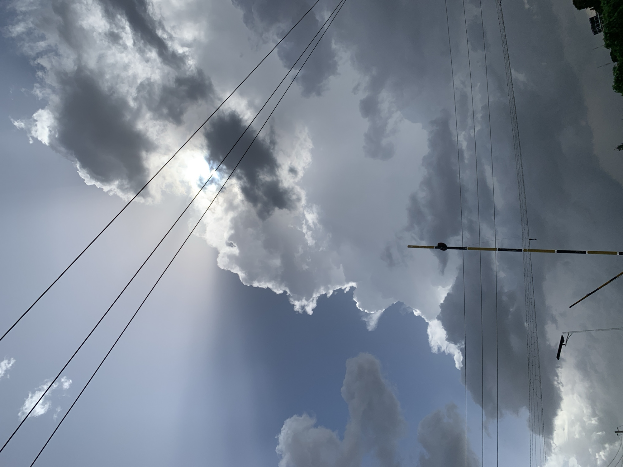 The media file shows a dramatic sky with a mix of dark, heavy clouds and bright patches where the sun is peeking through. The clouds create a sense of impending weather, possibly rain, but the sunlight suggests a break in the storm. Several power lines stretch across the frame, adding a man-made element to the natural scene. One pole with a yellow and black marking is visible, possibly a warning sign or part of a utility structure. The lighting is dynamic, with beams of light cutting through the darker clouds, creating high contrast. The overall impression is one of a powerful and changing sky, likely captured during the daytime, given the sunlight. The location context of Barakin Lerawi, Nigeria, suggests this is an outdoor scene, possibly in a semi-rural or urban environment where utility poles are common. There is no visible text in the image.