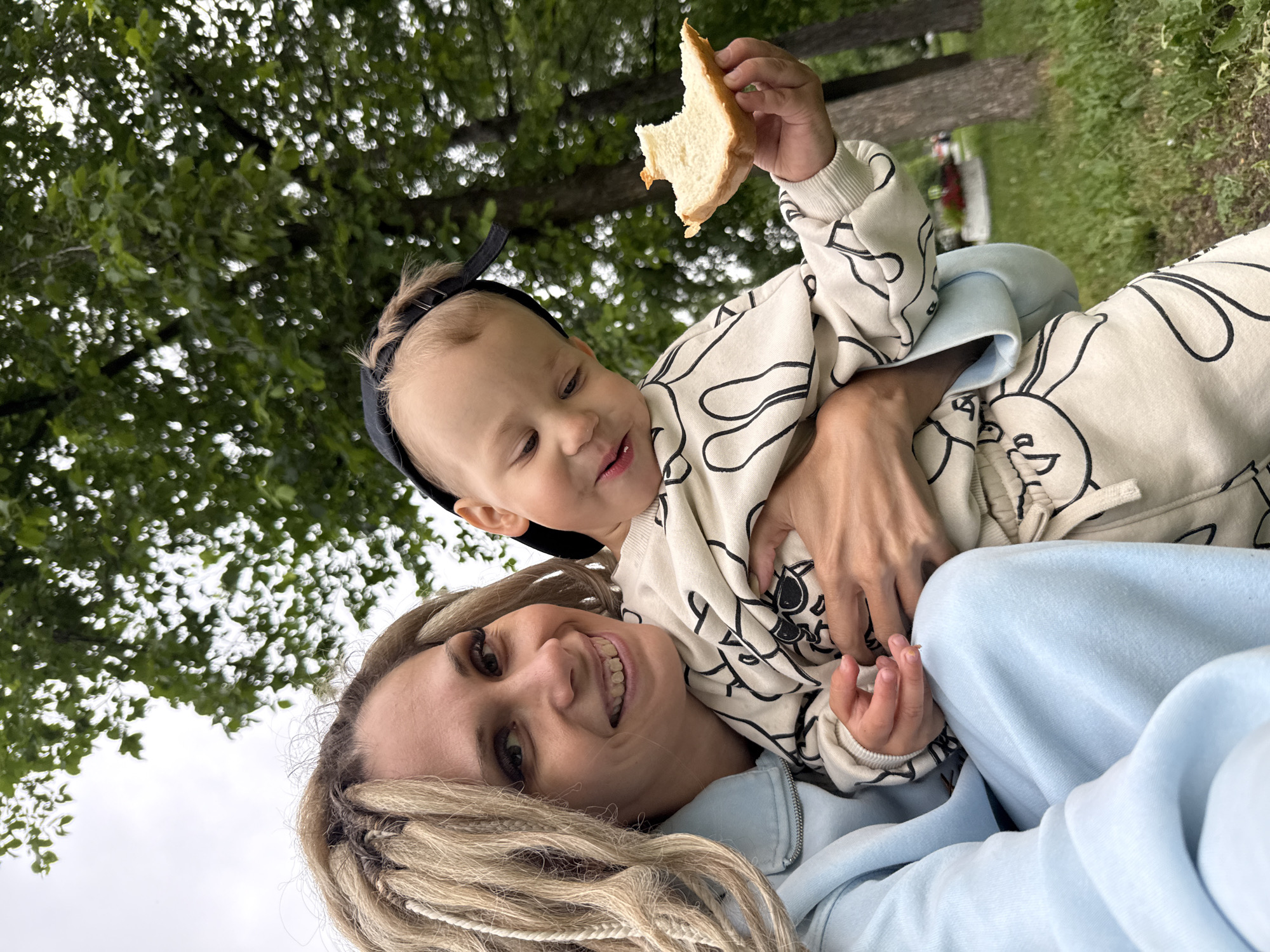 A light-skinned woman with blonde dreadlocks and bold eye makeup is smiling and holding a young child in her arms. She is wearing a light blue tracksuit. The child, wearing a light-colored sweatshirt with a black line drawing pattern and a black baseball cap, is looking down and holding a piece of white bread in his right hand. They are outdoors, with a background of green trees and grass, suggesting a park or outdoor recreational area. The sky appears overcast.