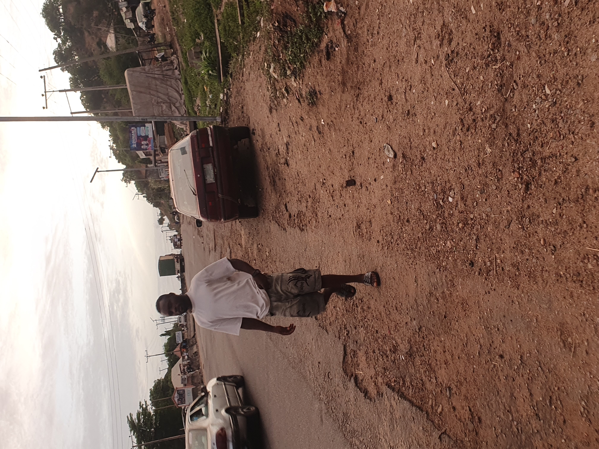 A street scene in Lokoja, Nigeria, shows a man walking along a partially paved road with a dirt shoulder. The man is wearing a white t-shirt and dark shorts. Several vehicles are visible, including a dark-colored sedan parked on the shoulder, a white sedan, and a truck in the distance. Utility poles and trees line the street. The sky is overcast.