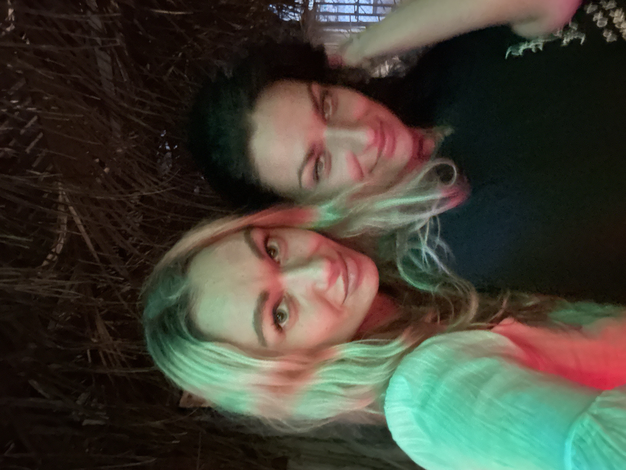Two women are featured in a close-up portrait, seemingly lying down or viewed from an overhead perspective. The woman in the foreground has long, wavy blonde hair and a light-colored top, while the woman behind her has darker hair and is wearing a black top. Both subjects are smiling and looking directly towards the camera, conveying a cheerful and relaxed mood.
The scene is dramatically illuminated by artificial colored lights, casting prominent red and green hues across their faces, hair, and clothing. This distinct lighting suggests a nighttime setting, possibly within a lively social environment like a club, bar, or event space. The background is dark and textured, appearing to be composed of dried, interwoven plant material such as straw or palm fronds, hinting at a natural or rustic setting. Given the location context of Mũi Né, Vietnam, this could be a beachside venue or a relaxed outdoor area common in a resort town. There is no visible text in the image.