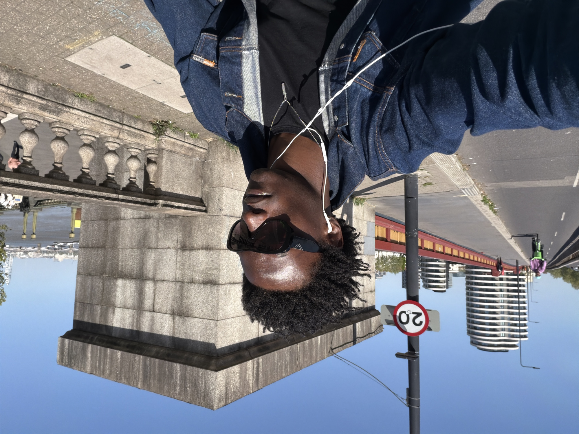 A man with dark skin and dreadlocks, wearing a black t-shirt and a blue denim jacket, takes a selfie on Tower Bridge in London, United Kingdom. He is wearing sunglasses and white headphones. The bridge's stone architecture and the River Thames are visible in the background, along with modern apartment buildings and a pedestrian walkway with a red railing. A "20" speed limit sign is also visible. Two cyclists are traveling on the bridge. The sky is blue.
