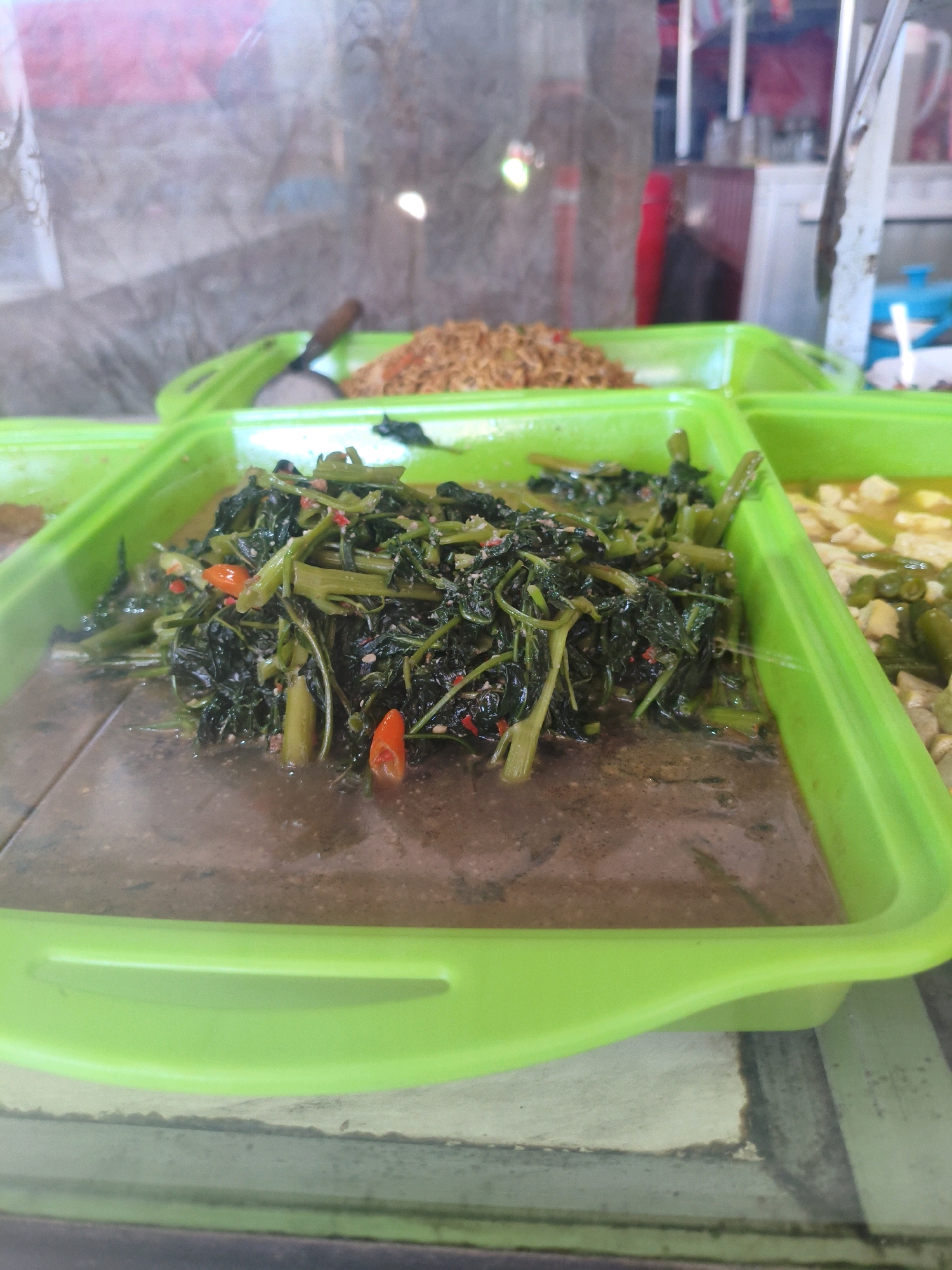 This image captures a close-up view of food displayed in a bright green plastic tray at an eatery in Bergas, Indonesia. The tray is segmented, with the most prominent section filled with a generous portion of stir-fried water spinach, also known as kangkung. The water spinach is cooked with what appears to be garlic, chilies, and a savory sauce, giving it a dark green and slightly glossy appearance. Several vibrant red chili peppers are visible amidst the greens, adding a pop of color and suggesting a spicy dish. The dish is served in a shallow pool of its cooking liquid.
Adjacent to the water spinach, in another segment of the tray, there is a dish of what seems to be tofu or tempeh in a light-colored broth with some green vegetables, possibly green beans, and perhaps some other ingredients like diced carrots. In a third, further-back section of the tray, a mound of what looks like fried rice or another grain-based dish is visible.
The food is presented behind a glass or transparent barrier, suggesting it is a display for customers to choose from, typical of a warung or local Indonesian restaurant. The background is blurred but hints at the interior of the establishment, with shelving and kitchen equipment visible. The overall lighting is bright, illuminating the food and the green tray. The style of the food presentation is casual and indicative of local Indonesian cuisine.