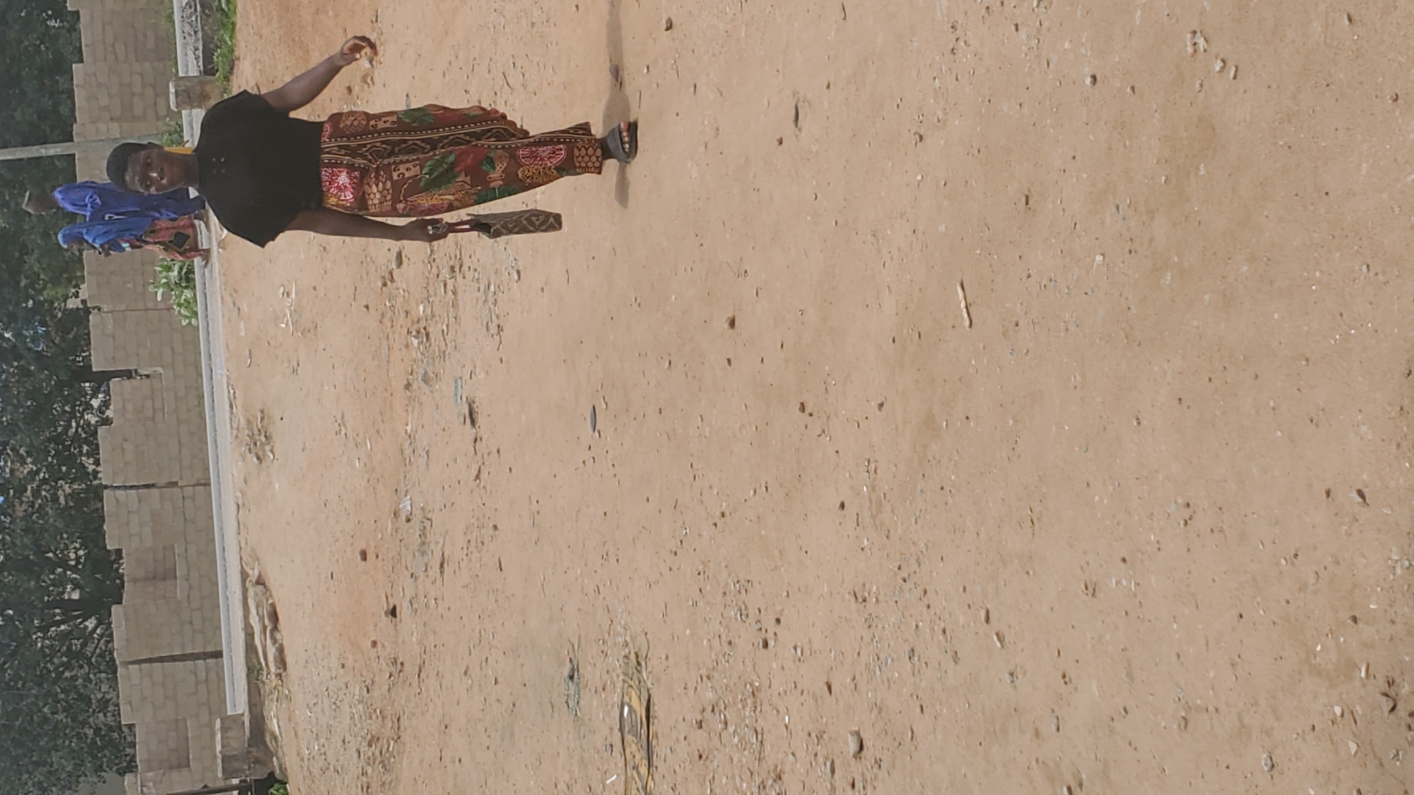 The image shows a person walking along a dirt road. The person is wearing a black top and a colorful skirt. There are two other people in the background, also walking. The scene appears to be in a rural or semi-rural area, with some brick structures and trees visible on the left side of the image. The ground is sandy and dry, suggesting a warm, possibly sunny day. The lighting suggests it is daytime, but the exact time is unclear. There is no visible text in the image.