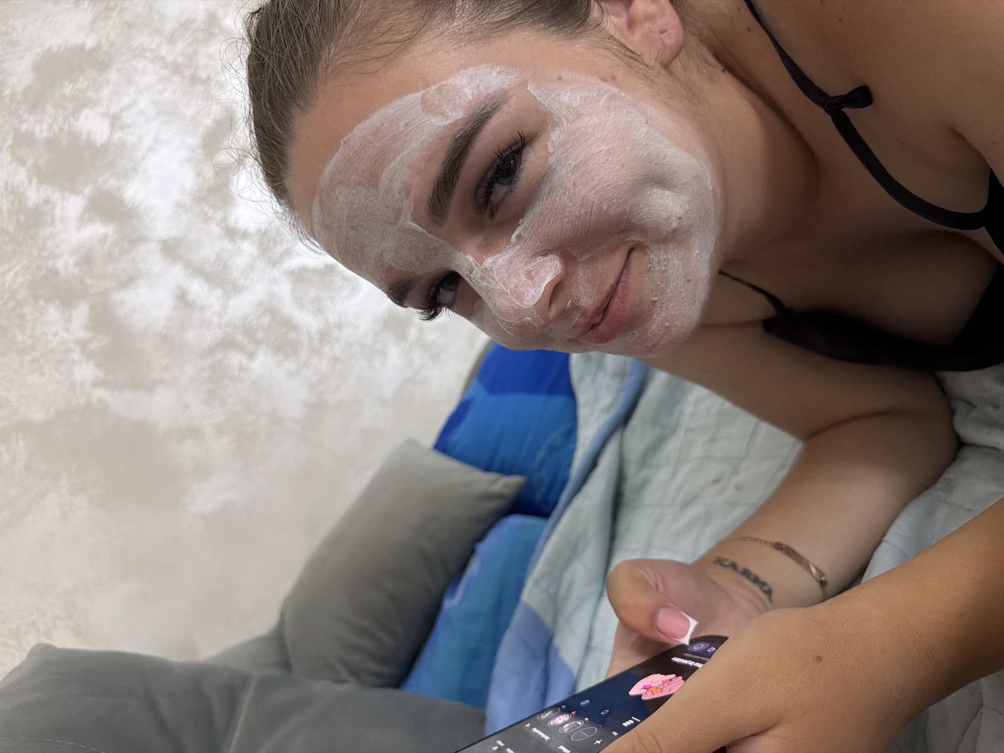 A young woman with a white facial mask applied is lying on a bed, holding a smartphone in her left hand. She is looking at the phone with her head slightly tilted. The room appears to be a bedroom, with pillows and bedding visible. The background shows a textured wall with a subtle pattern. The lighting is soft and natural. The image does not provide any information about the location in the city of Strășeni, Moldova.