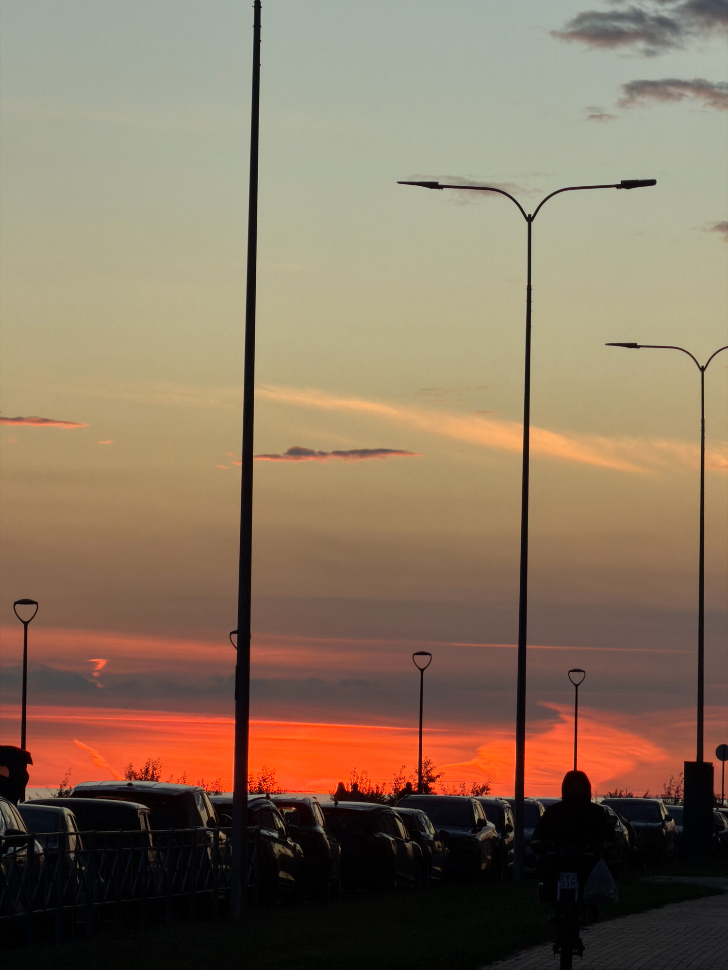 In the foreground, a person rides a scooter on a paved path. The person is silhouetted against a vibrant sunset sky, wearing a dark hooded jacket and carrying a bag. Behind the person, a row of parked cars is visible, silhouetted against the fiery orange and pink hues of the sunset. Tall poles, likely streetlights, are scattered throughout the scene, some casting long shadows. The sky is filled with dramatic clouds, illuminated by the setting sun. The scene appears to be a parking lot or an area adjacent to a road, possibly on the outskirts of Saint Petersburg, Russia, given the architectural style of the streetlights and the overall atmosphere. The overall impression is one of urban twilight, with the natural beauty of the sunset contrasting with the man-made structures.