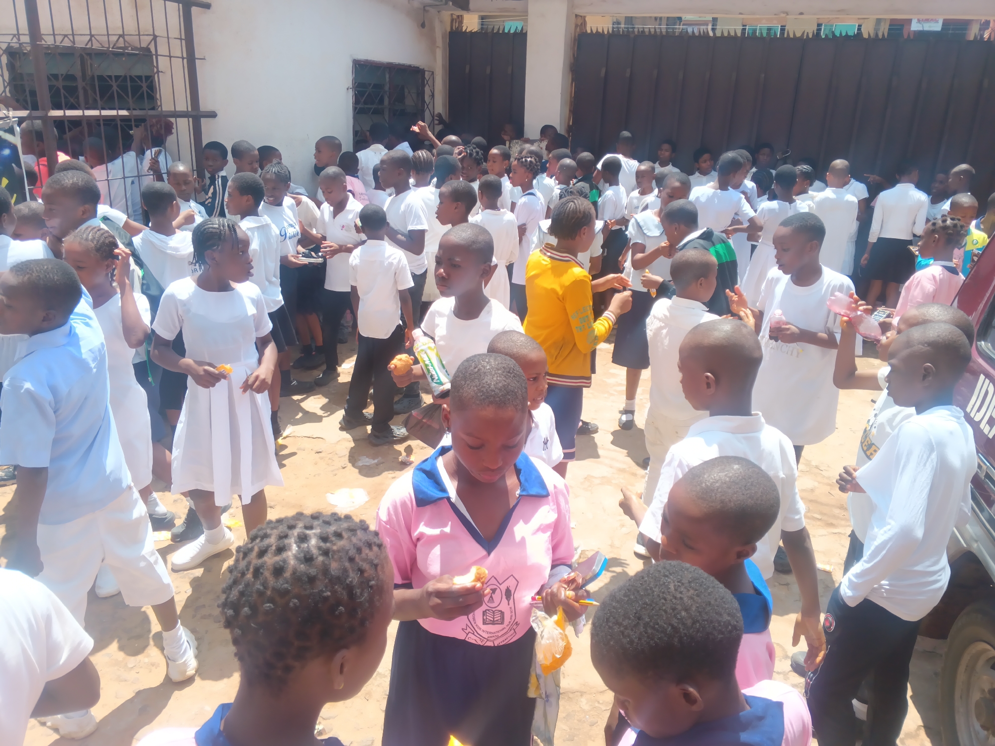 This photograph captures a gathering of children, likely students, outside a building with a metal gate and a dark, corrugated metal fence. The scene appears to be a school or community event. The children, predominantly dressed in white or light-colored school uniforms, are engaged in various activities. Many are standing in groups, some holding food items and plastic bottles, suggesting a snack or refreshment break.
One girl in the foreground, wearing a pink and blue collared shirt and a dark skirt, is holding a piece of food and a plastic bag. Her gaze is directed downwards, as if examining the food. To her left, another child with braided hair is also looking at something in their hands.
The time of day is likely daytime, with bright sunlight casting shadows on the sandy ground. The weather appears to be warm and sunny. The overall mood seems to be one of casual interaction and enjoyment among the children.
Onitsha, Nigeria, is provided as a location context. While not directly visible in the image, this context suggests a likely tropical climate.
Visible text includes a logo on the pink shirt of the girl in the foreground. The logo contains a crest with what appears to be an open book and a laurel wreath, along with text that includes "GLOBAL SCHOOL" and "AWARD." This confirms the presence of a school setting. Another child in a white shirt has a partially visible logo that reads "CITY."