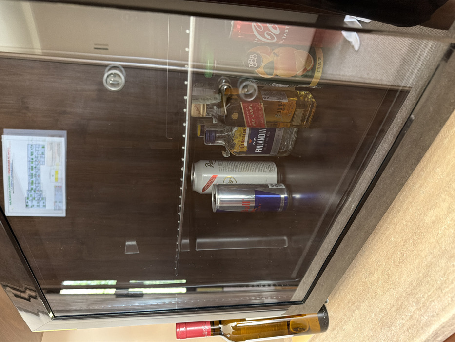 The image displays an interior scene, likely a hotel room or similar accommodation in Kladnitsa, Bulgaria. A compact, dark-colored mini-fridge with a transparent glass door is centrally positioned on a light brown wooden or laminate countertop. Inside the mini-fridge, from left to right, are: one silver and blue Red Bull energy drink can, one tall white can possibly labeled "Radler," one small clear bottle of Finlandia vodka with a blue label, one small amber-liquid bottle of Johnnie Walker Red Label Scotch whisky, one orange-themed can of "BBB Orange" soda, and one red Coca-Cola can. To the immediate left of the mini-fridge, on the countertop, stands a tall, clear glass bottle with a red foil cap, containing a light amber liquid consistent with white wine. Above the mini-fridge, a document enclosed in a clear protective sleeve is taped to the dark wooden cabinet. This document features a schematic floor plan with green-highlighted sections and Cyrillic text, including phrases such as "ПОЖАРНА БЕЗОПАСНОСТ" (FIRE SAFETY) and "НАПЪЛНО ЗАБРАНЕНО ПУШЕНЕТО НА ОТКРИТ ОГЪН" (SMOKING AND OPEN FLAMES ARE STRICTLY FORBIDDEN). The glass door of the mini-fridge reflects vertical light streaks, a light-colored carpeted floor, and a dark object or shadow on the right side of the frame. No individuals are present.