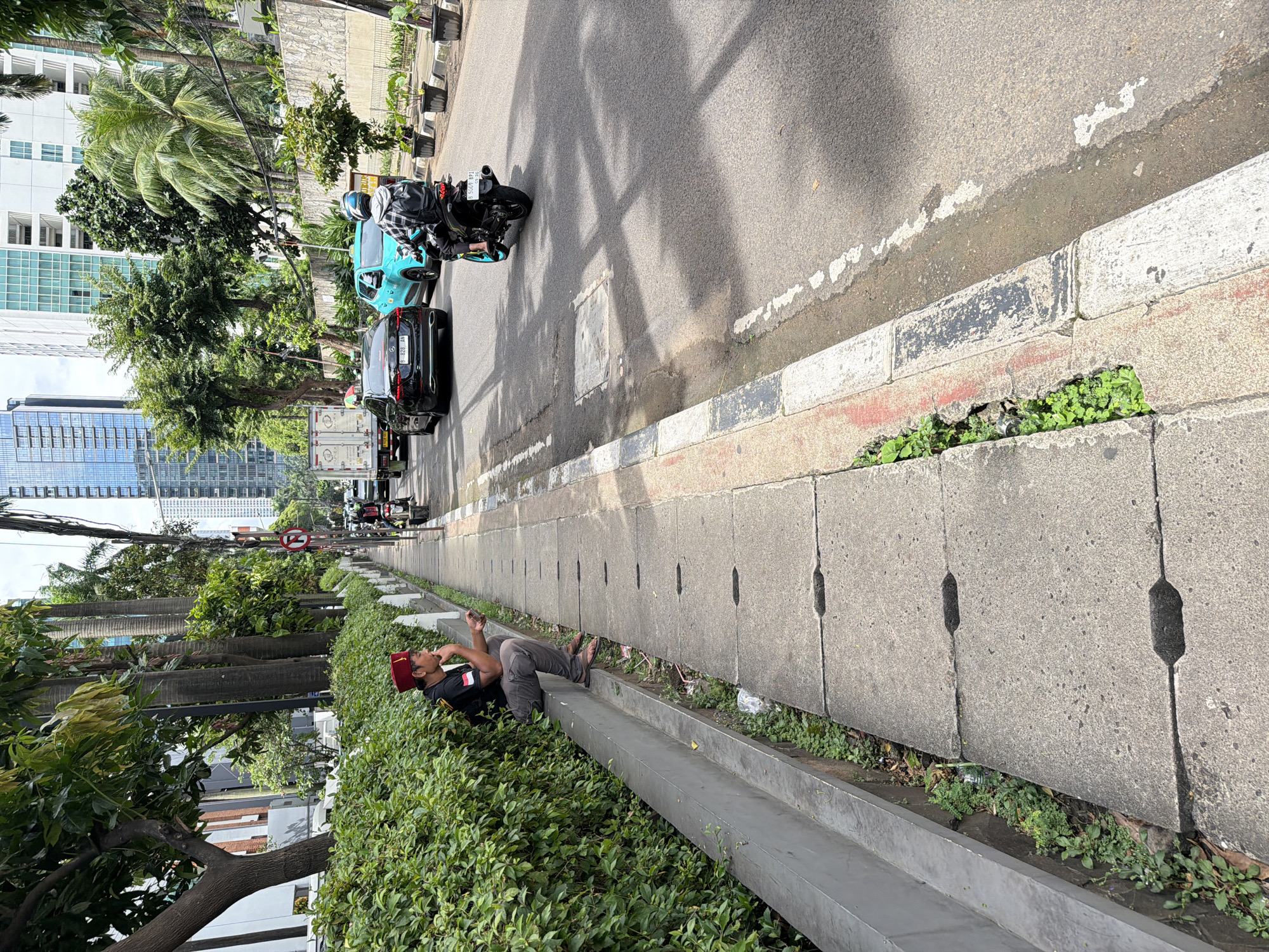 The image captures an urban scene in South Jakarta, Indonesia. A man wearing a red fez and a black shirt with an Indonesian flag patch is lying on his back, seemingly using his phone. He is positioned beside a concrete drainage channel, with lush green bushes surrounding him. Behind him, a street is visible, featuring traffic, including a motorcycle and cars. Skyscrapers and tall buildings form the backdrop, suggesting a densely populated urban environment. The photo is taken at an angle, tilting the perspective of the street and buildings.
