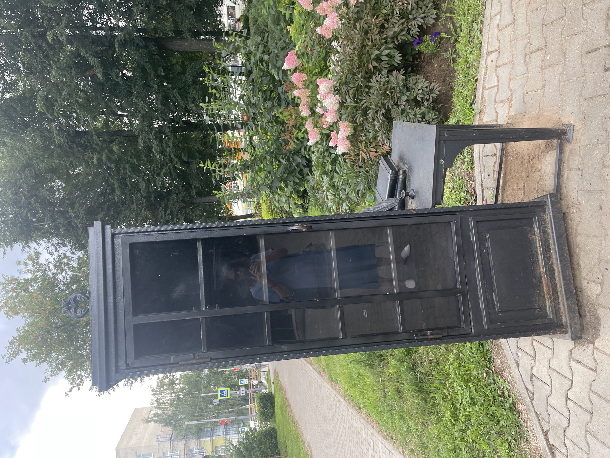 A tall, black, narrow cabinet with glass panes stands tilted on a paved walkway next to a street with some vegetation. The cabinet has a decorative metal element on top and a more solid panel at the bottom. A small, black metal desk is attached to the right side of the cabinet, holding two books and an inkwell with a quill. A person in a blue dress is reflected in the glass of the cabinet, holding a phone and taking a selfie. In the background, there are trees, bushes with pink flowers, a building under construction, and street signs. The scene appears to be in a public urban area in Chaikovsky, Russia, possibly a park or a street.