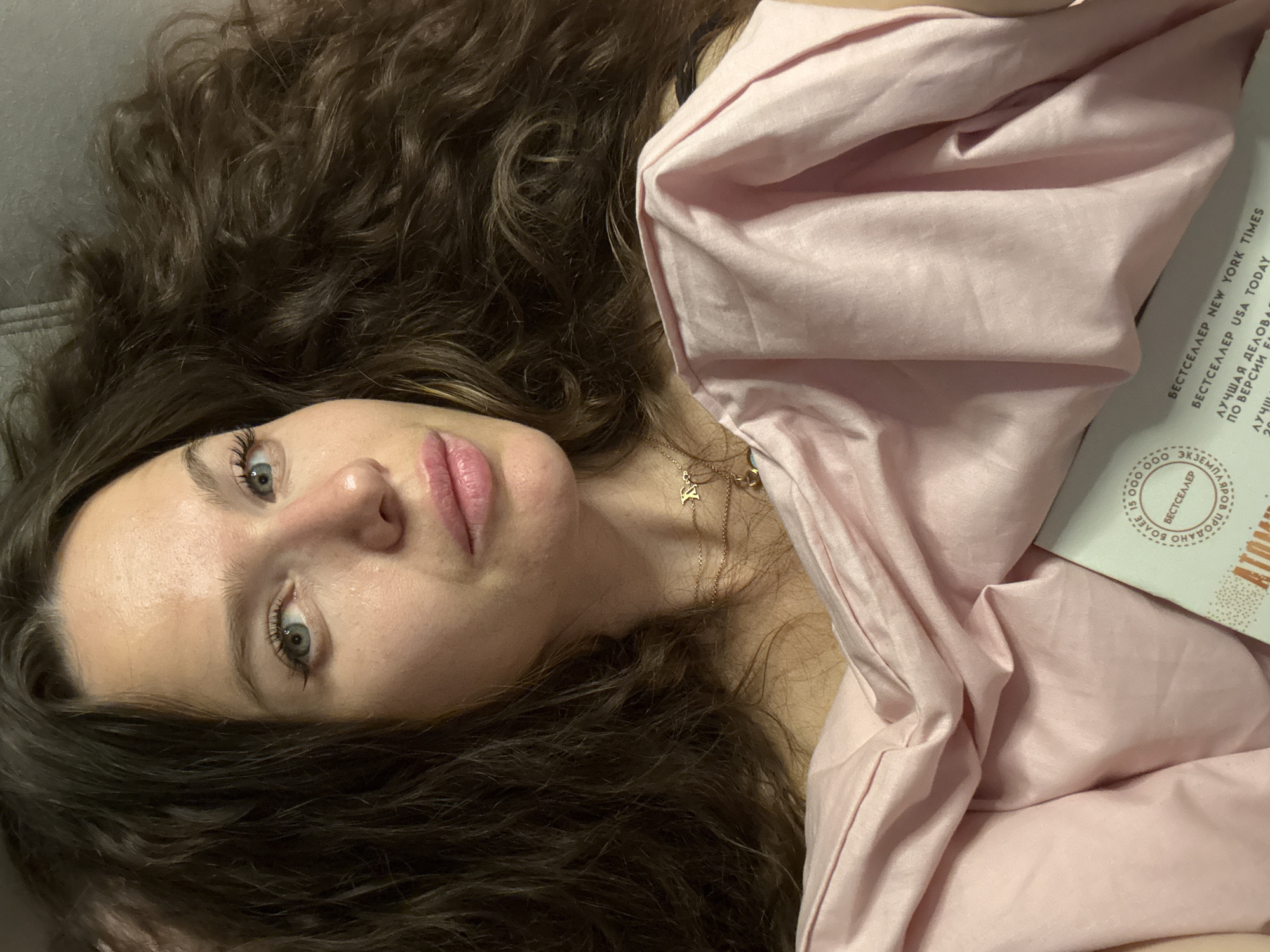 A high-angle portrait captures a woman with long, voluminous dark brown curly hair lying down, looking directly at the camera. Her head rests on a dark grey surface. She has striking light blue-grey eyes, a fair complexion, and full, slightly parted lips with a pink, glossy finish. Her gaze is direct and calm.
She wears a delicate gold-colored necklace with a small pendant. Her upper body is partially covered by a soft, light pink fabric, possibly silk or satin, draped around her. To her right, a white book or magazine cover is partially visible, featuring Cyrillic text.
The visible text indicates the item is a "БЕСТСЕЛЛЕР" (Bestseller), specifically mentioning "NEW YORK TIMES" and "USA TODAY" bestsellers. It also notes "БОЛЕЕ 15 000 000" (More than 15,000,000) copies sold and phrases like "ЛУЧШАЯ ДЕЛОВА" (Best Business). A partial logo containing "АТОМ" (Atom) is discernible, suggesting it may be the book "Atomic Habits." The Russian text is consistent with the location context of Khimki, Russia. The image appears to be taken indoors, emphasizing a relaxed and intimate atmosphere.