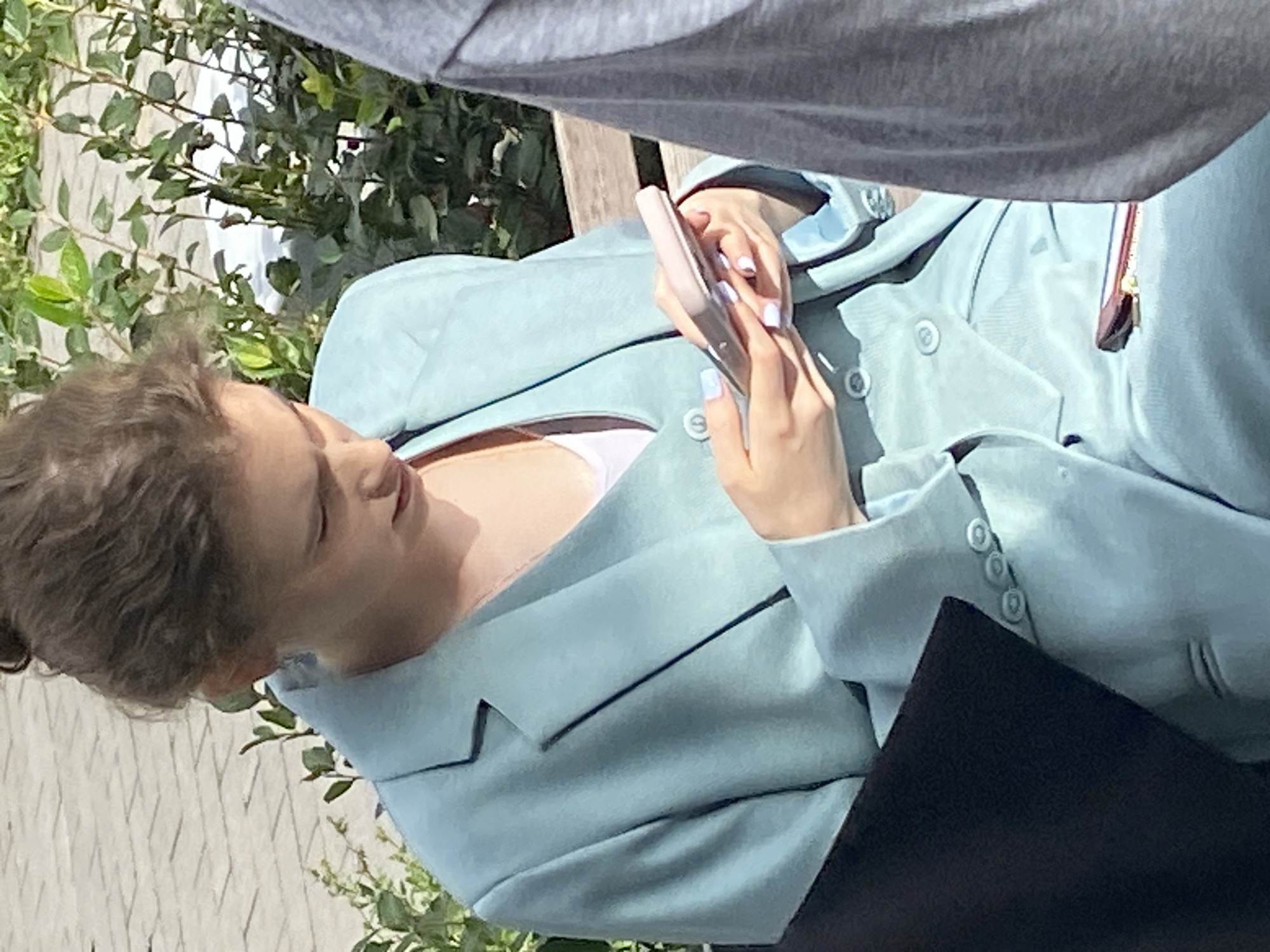 A young woman with her hair in a bun is sitting on a park bench outdoors, looking down at her phone. She is wearing a light blue suit with a matching vest and white undershirt. Her fingernails are painted a pale lavender. The background shows green foliage and a paved area. A person in a gray shirt is partially visible on the right side of the frame. There is a dark object on the bench to the left of the woman.