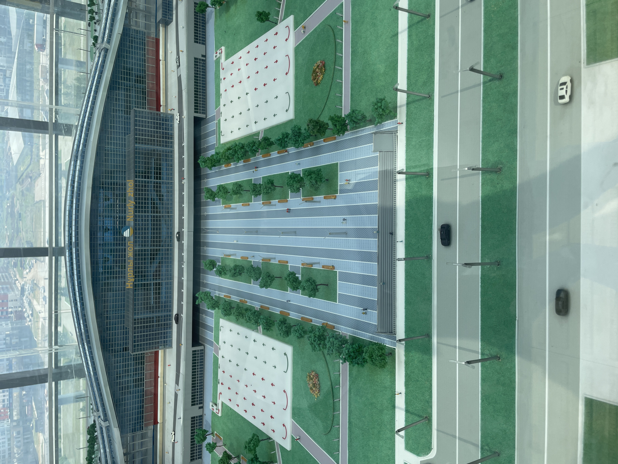 This is a high-angle shot of a detailed architectural model of Nurly Zhol railway station in Astana, Kazakhstan. The model showcases the station's modern, curved glass facade with the station's name prominently displayed in yellow lettering. In front of the station is a vast, open plaza with marked pedestrian walkways, rows of artificial trees, and benches. On the plaza, there are several small figures representing people. To the sides of the plaza are two large, rectangular white platforms with numerous red and green arrows indicating traffic flow. Beyond the plaza, a road with several miniature cars and lamp posts is depicted. The model is positioned indoors, as evidenced by the reflection of a window in the foreground and the distant cityscape visible through the window behind the model.