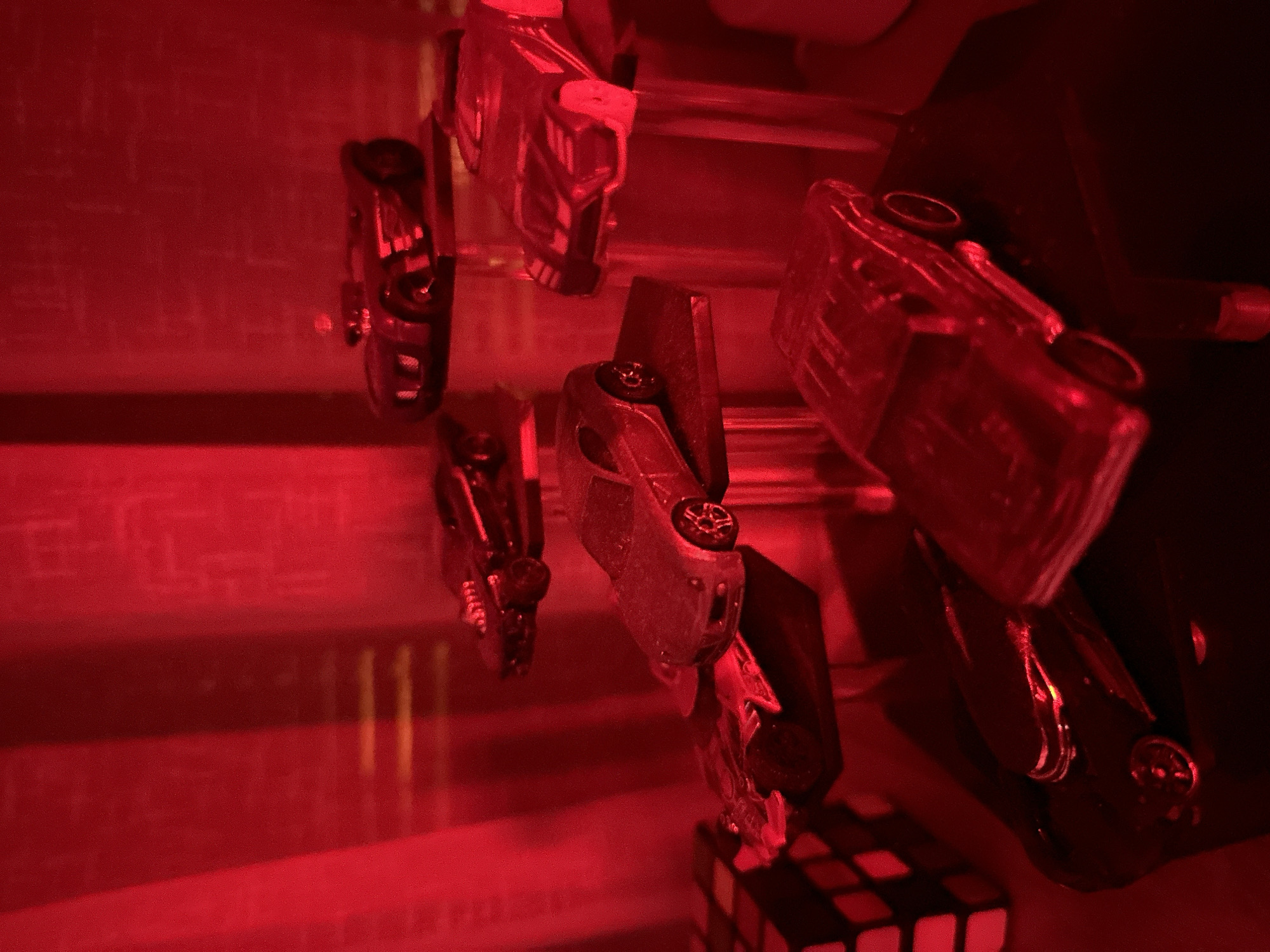 A collection of toy cars is displayed on various levels. In the foreground, a black toy car is positioned on a dark surface. Behind it, a transparent or light-colored toy car with visible wheels sits on a similar surface. To the left, a set of three colored cubes, red and black, is visible.
Further back and slightly to the left, a silver toy car with detailed wheels is mounted on a clear stand. Another toy car, dark colored with a prominent grill and exhaust pipes, rests on a raised platform behind the silver car.
In the upper right quadrant, a black toy car with a streamlined design and visible engine details is displayed on a dark stand. Next to it, to the right, is a silver toy car with a distinct grill and headlights, also on a clear stand.
The background consists of vertical red blinds or curtains, creating a dramatic lighting effect that casts a red hue over the entire scene. The lighting suggests an indoor setting, possibly a display or shelf arrangement, in an unknown location.
