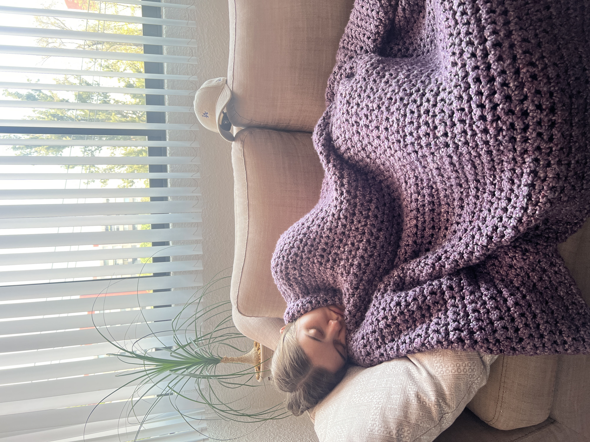 A woman is sleeping on a tan sofa covered with a large purple crochet blanket. Her head rests on a decorative pillow. Beside the sofa, near a window with closed blinds, is a small potted plant. A baseball cap with the letter "LA" is perched on the back of the sofa. The location is not specified but is possibly in Mexico City.