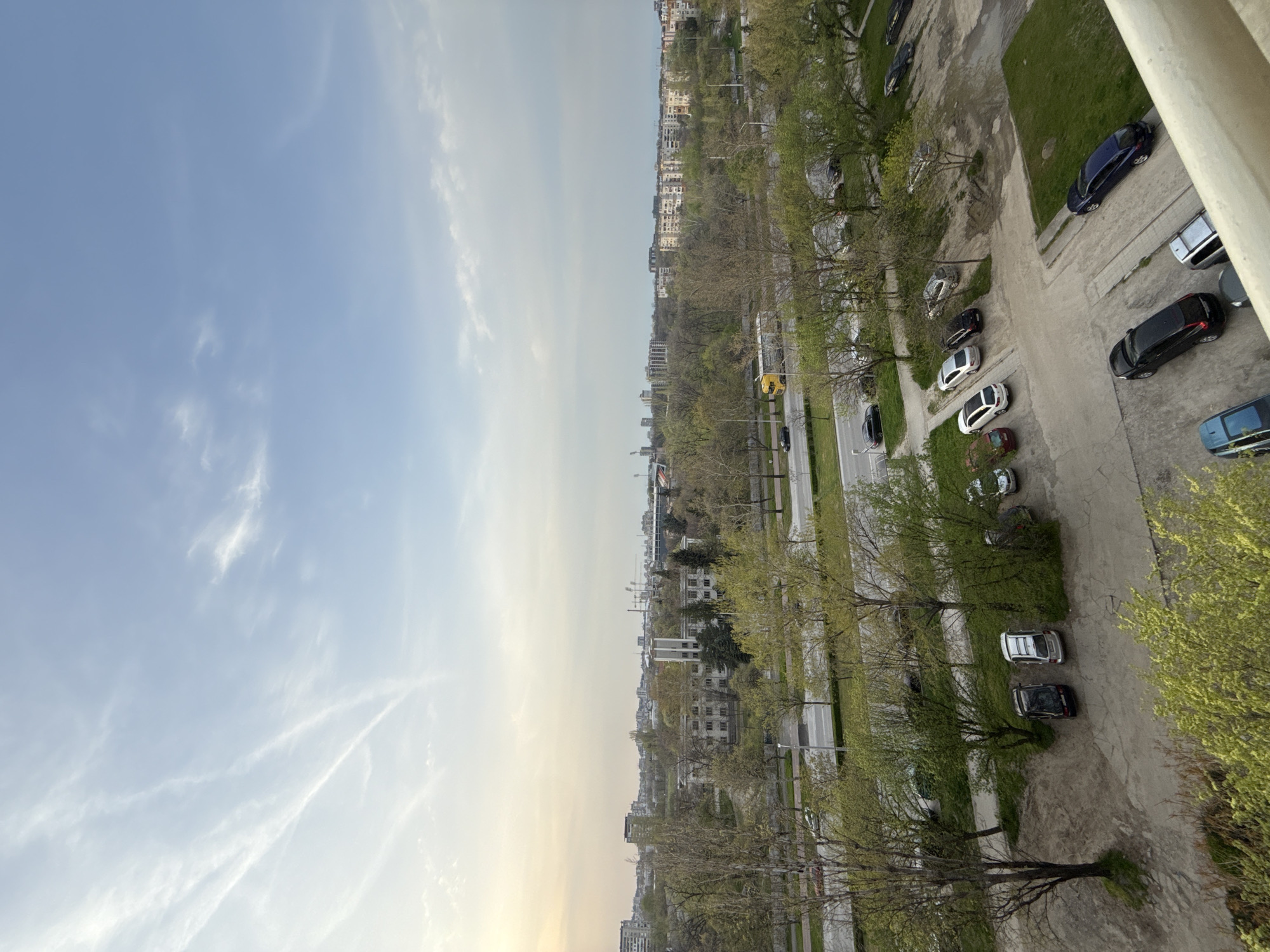 The image shows an elevated view of a cityscape, likely taken from a high-rise building. The sky is a gradient of light blue to a faint yellow near the horizon with wispy cirrus clouds. The central part of the view is dominated by rows of buildings interspersed with trees. A main thoroughfare runs through the middle of the frame, with vehicles visible. Adjacent to the roadway is a parking area with numerous parked cars. The ground below the trees appears to be a mix of paved and unpaved surfaces, with areas of green grass. Based on the architecture, vegetation, and general layout, the location is plausibly in Пловдив, Bulgaria, particularly the area around the Maritza River and the city's parks, though without additional context, this identification is tentative.