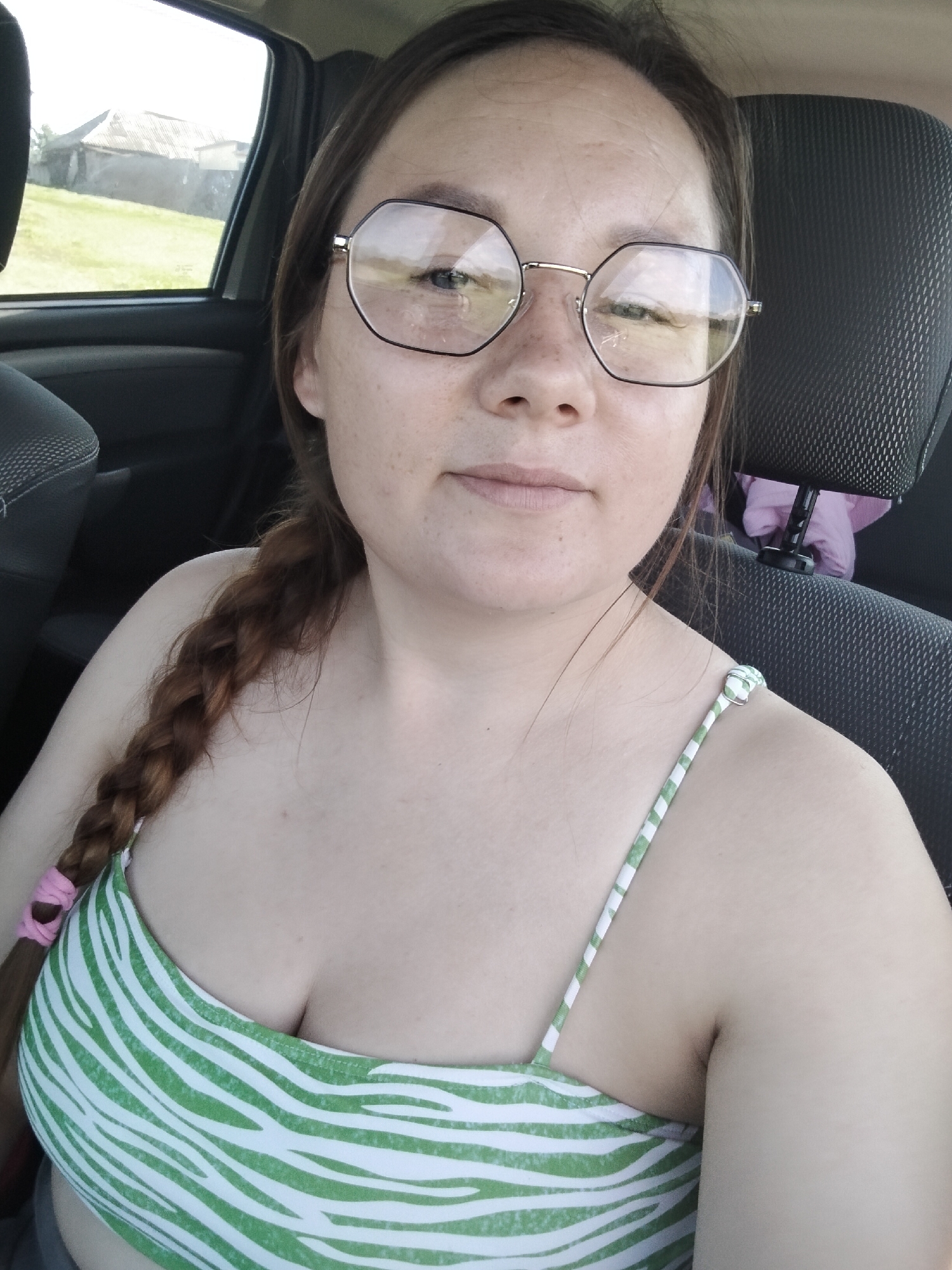 A woman with brown hair in a braid is sitting in the passenger seat of a car. She is wearing glasses with large, hexagonal frames and a green and white striped top. The woman is looking at the camera. The background shows a glimpse of the exterior through the car window, with a building and greenery visible under a bright sky, suggesting it is daytime and likely good weather. The scene appears to be a rural or semi-rural setting in Блохино, Russia. The woman's expression is neutral. There is no visible text in the image.
