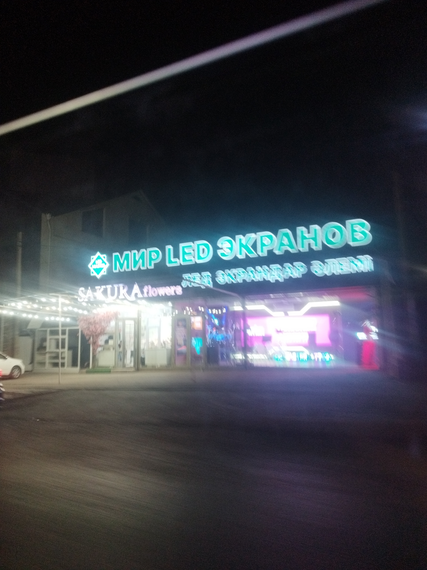 The image shows a storefront at night in Shymkent, Kazakhstan. The storefront is illuminated by bright LED signs. The main sign reads "МИР LED ЭКРАНОВ" in large, green, block letters. Below this sign, another sign, also illuminated, reads "SAKURA flowers". Further to the right, another sign is visible with what appears to be text in Cyrillic script. The building has a modern facade with large windows. Inside, the lighting is bright and colorful, with what look like displays or products visible. A stylized pink tree is located near the entrance. A white car is parked on the left side of the image. The street in front of the building is asphalt. The overall impression is of a commercial establishment operating at night.