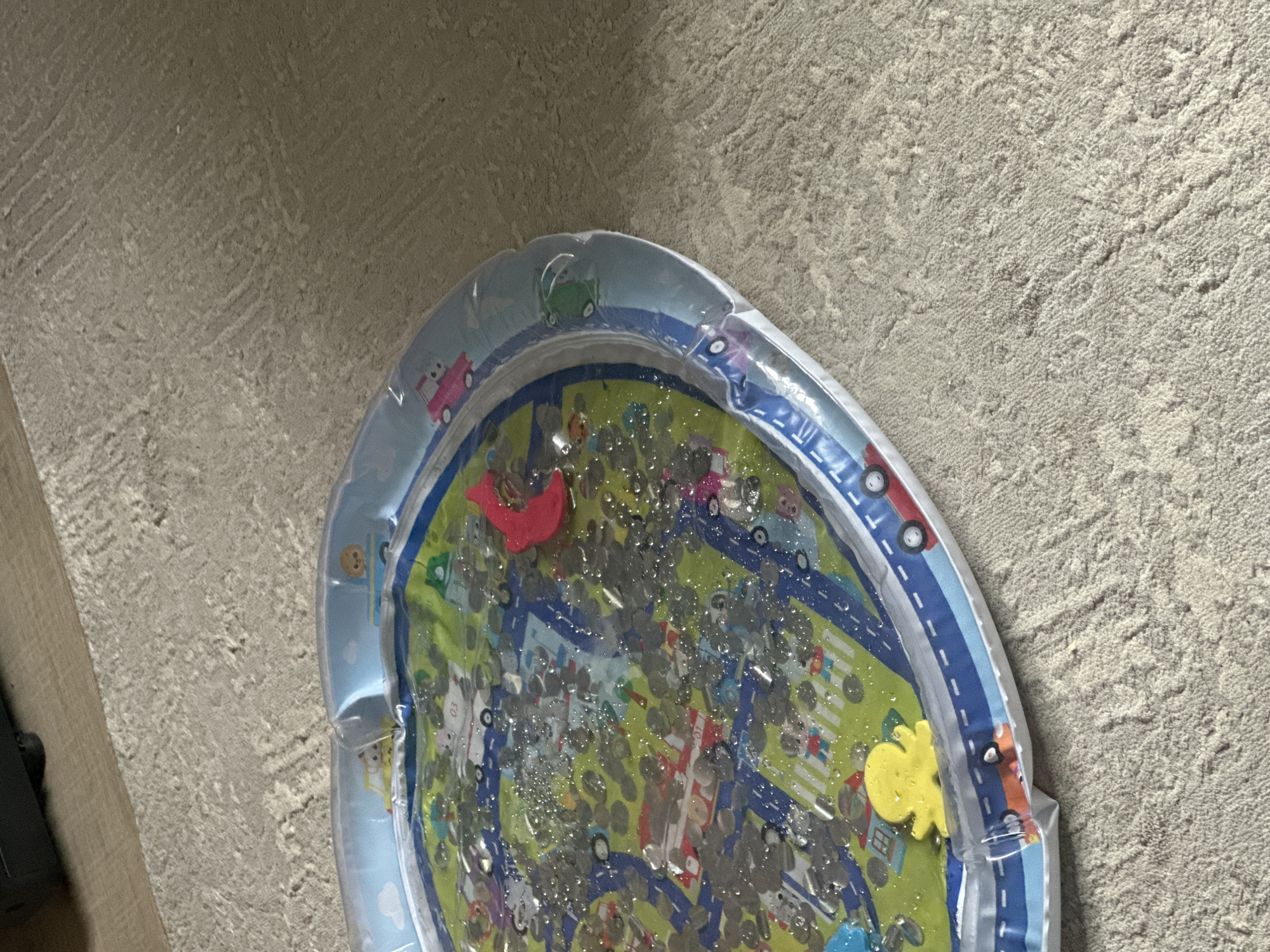 The image shows an inflatable water mat for babies lying on a textured carpet. The mat has a colorful design featuring a road with toy cars and houses, and it is filled with water and small floating objects that appear to be sequins or glitter. A bright yellow rubber ducky and a pink toy are also visible on the mat. The mat is partially deflated around the edges. The background shows a wooden surface and the carpeted floor. The location is Pervouralsk, Russia.