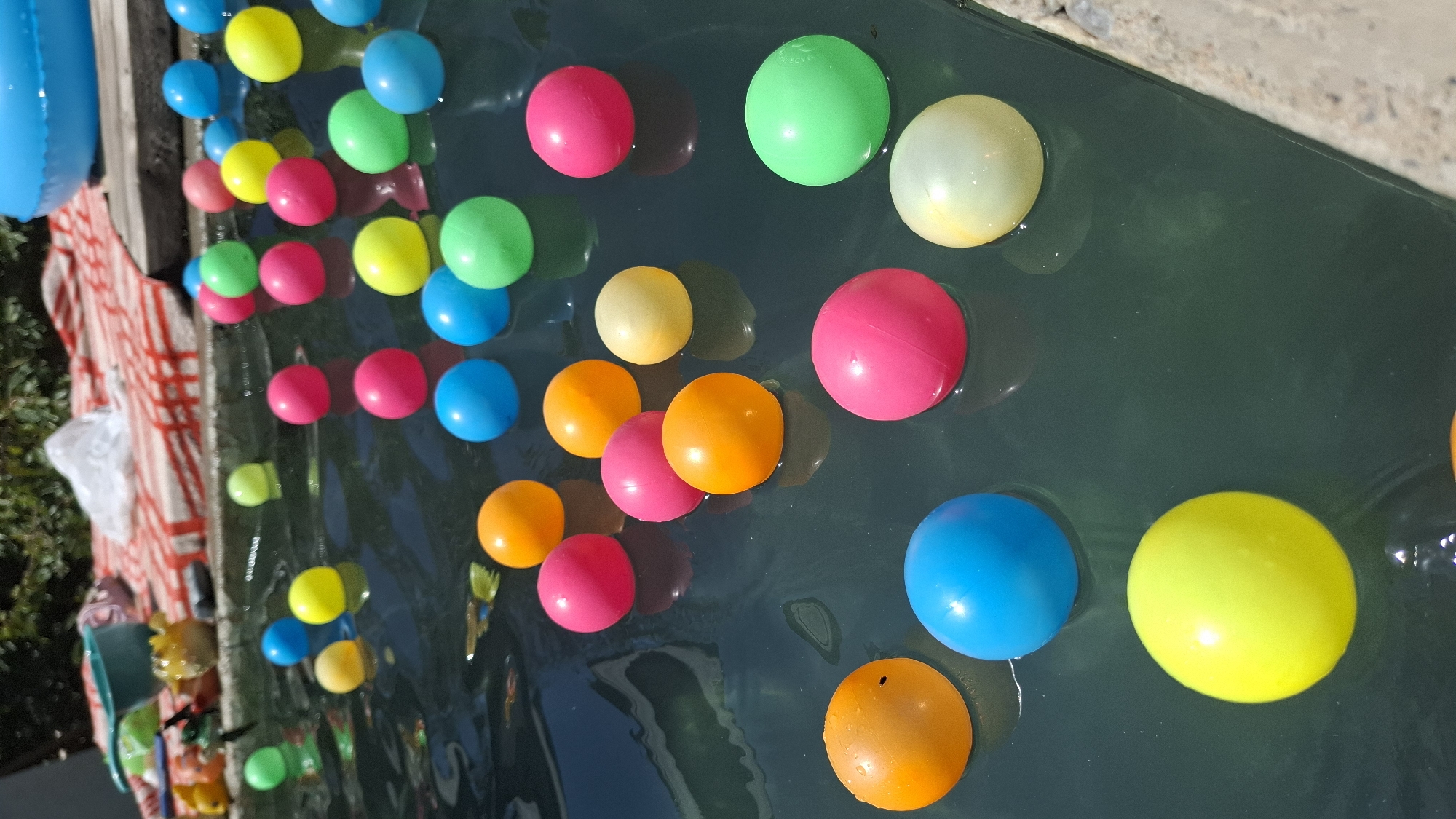 This image depicts a close-up, overhead view of numerous colorful, spherical balls floating on the surface of dark, slightly rippling water. The balls are predominantly pink, orange, yellow, blue, and green, with a few appearing lighter in color, possibly white or pale yellow. They are scattered across the frame in varying densities, with some clusters and some more isolated.
In the background, partially visible, is a pool edge or structure made of concrete, with some textured tiles or stones. Beyond that, there is a red and white striped cloth or towel laid out, and some out-of-focus green foliage suggests an outdoor setting, possibly a garden or yard. A blue inflatable ring is also visible in the upper right corner, hinting at a recreational water area. There are no people or animals clearly visible in the image. The scene appears to be set in an outdoor swimming or wading pool, likely during daylight hours, given the lighting and shadows. The location is not identifiable as Шұбарсу, Kazakhstan based on the visual information provided.