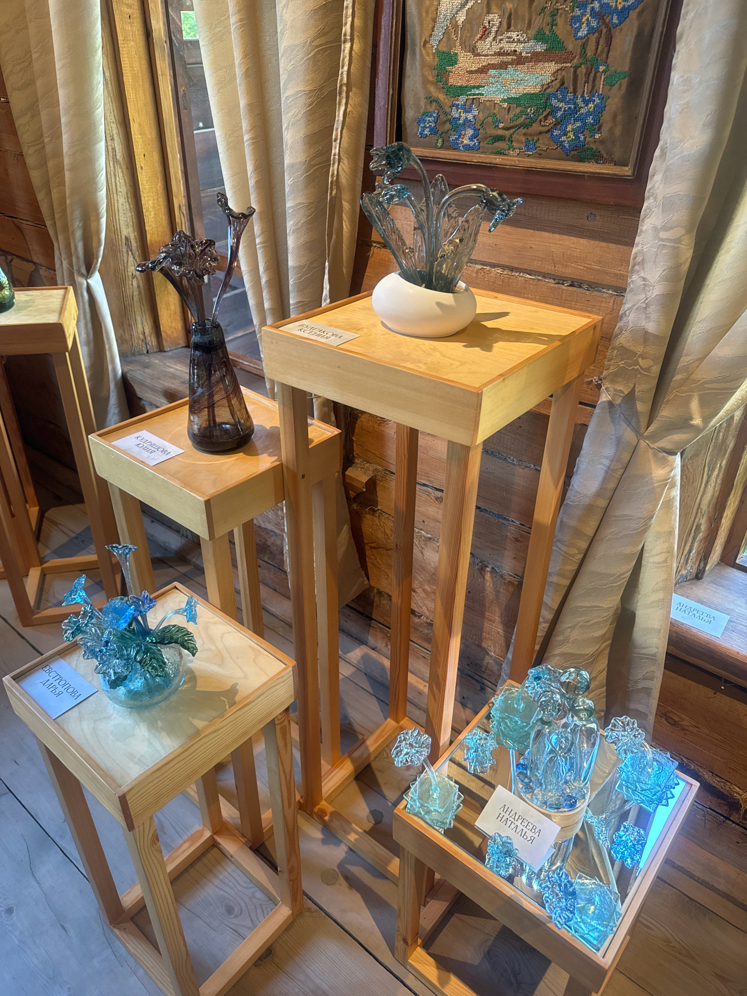 The image captures an indoor exhibit featuring glass art pieces displayed on wooden stands within the Тальцы museum complex in Irkutsk, Russia. The walls are paneled with rough-hewn logs, characteristic of traditional Siberian architecture. Beige curtains hang behind the display, adding a soft texture to the rustic setting.
On a taller wooden stand in the center, a glass sculpture of stylized flowers in muted tones is presented in a white ceramic bowl. A label reads "БУЛДАКОВА КСЕНИЯ". To the left, a smaller wooden stand holds a dark, swirling glass vase. A label on this stand states "КУДРЯШОВА ЮЛИЯ".
In the foreground to the left, a lower, squarish wooden stand displays a clear blue glass sculpture of flowers in a clear bowl. The label on this stand reads "ЕВСТРОПОВА ДАРЬЯ".
To the right, on another low wooden stand with a mirrored surface, is a collection of various light blue and clear glass art pieces, including what appears to be a couple embracing and abstract floral shapes. A label on this stand says "АНДРЕЕВА НАТАЛЬЯ". The wooden floor is visible beneath the stands. An embroidered artwork depicting a swan and blue flowers is mounted on the log wall in the upper right portion of the image. The overall scene suggests a craft or art exhibition in a museum setting.
