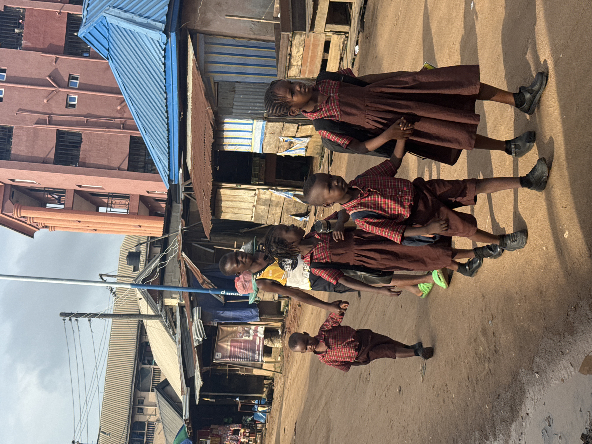 The image shows a group of six children, primarily in school uniforms, walking along a dirt road in what appears to be a residential or commercial area. Most children wear plaid shirts (red, black, and white pattern) with dark brown shorts or dresses, and black shoes, suggesting they are either heading to or from school. Several of them carry backpacks and some are holding hands. An older individual wearing a yellow, white, and black patterned t-shirt is partially visible walking alongside them.
The setting features an unpaved, dusty road. To the left, a multi-story reddish-brown building with multiple windows is visible, partially obscured by a bright blue corrugated metal roof. In the background and to the right, there are numerous informal structures with corrugated metal roofs and walls, likely houses or small shops. Utility poles and a tangle of electrical wires are present against a bright, slightly overcast sky.
The strong sunlight creates long, distinct shadows on the ground, indicative of either early morning or late afternoon. The children have neutral expressions, with some looking towards the camera. Text "NDDO" is visible on the older individual's t-shirt. A banner in the background has some illegible text. The scene captures a moment of daily life in a bustling community, likely in Ihiagwa, Nigeria, given the context.