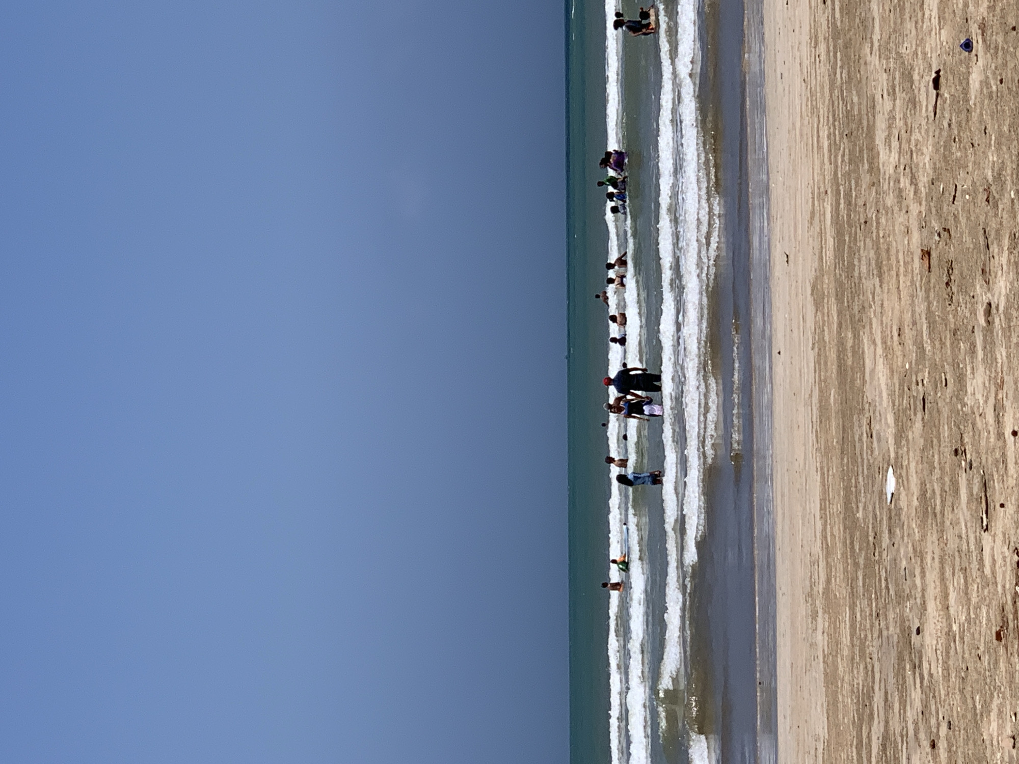 The image depicts a scene at Weligama Beach, Sri Lanka. The ocean is active with moderate waves, and several people are enjoying the water. In the foreground, a sandy beach with scattered debris is visible. The middle ground shows the ocean with numerous individuals wading, swimming, and standing in the waves. Some are interacting with each other, while others are enjoying the water individually. The background is dominated by a vast expanse of blue sky meeting the ocean horizon. The lighting suggests it is daytime, likely with bright sunlight.