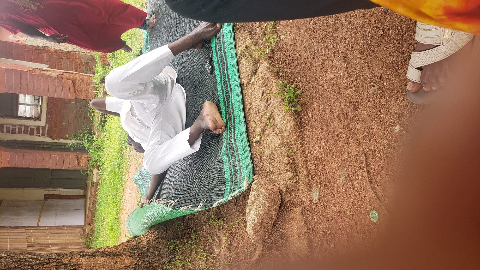 A dark-skinned man wearing white clothing is lying on his back on a green and black mat outdoors. He is positioned on the ground next to a tree trunk. His head is propped up by his hands, and his legs are extended. In the background, a red brick building with arched doorways and a broken window is visible. Green grass and red soil make up the surrounding environment. To the right, a person wearing a red garment and another in black clothing are partially visible. A few small green plants are growing in the dirt.