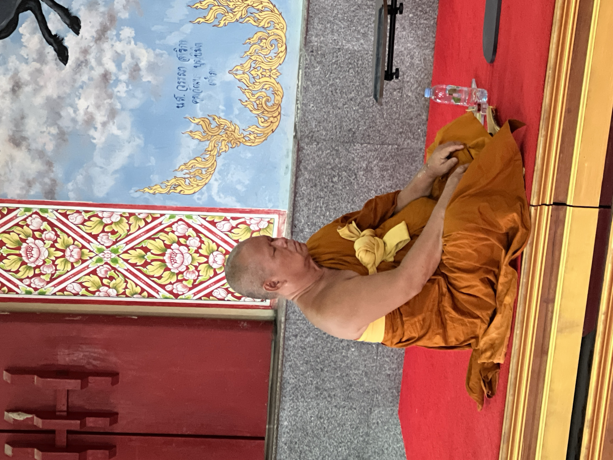 A Buddhist monk in an orange robe sits cross-legged on a red carpeted platform in Baan Plai Laem, Thailand. His eyes are closed, and his head is bowed, suggesting meditation. To his right, a clear plastic water bottle stands next to a small glass container with a red substance. The monk is positioned in front of a wall decorated with traditional Thai floral patterns and a mural of a horse in the sky. To the left, a red wooden structure is visible. The overall scene is peaceful and spiritual.