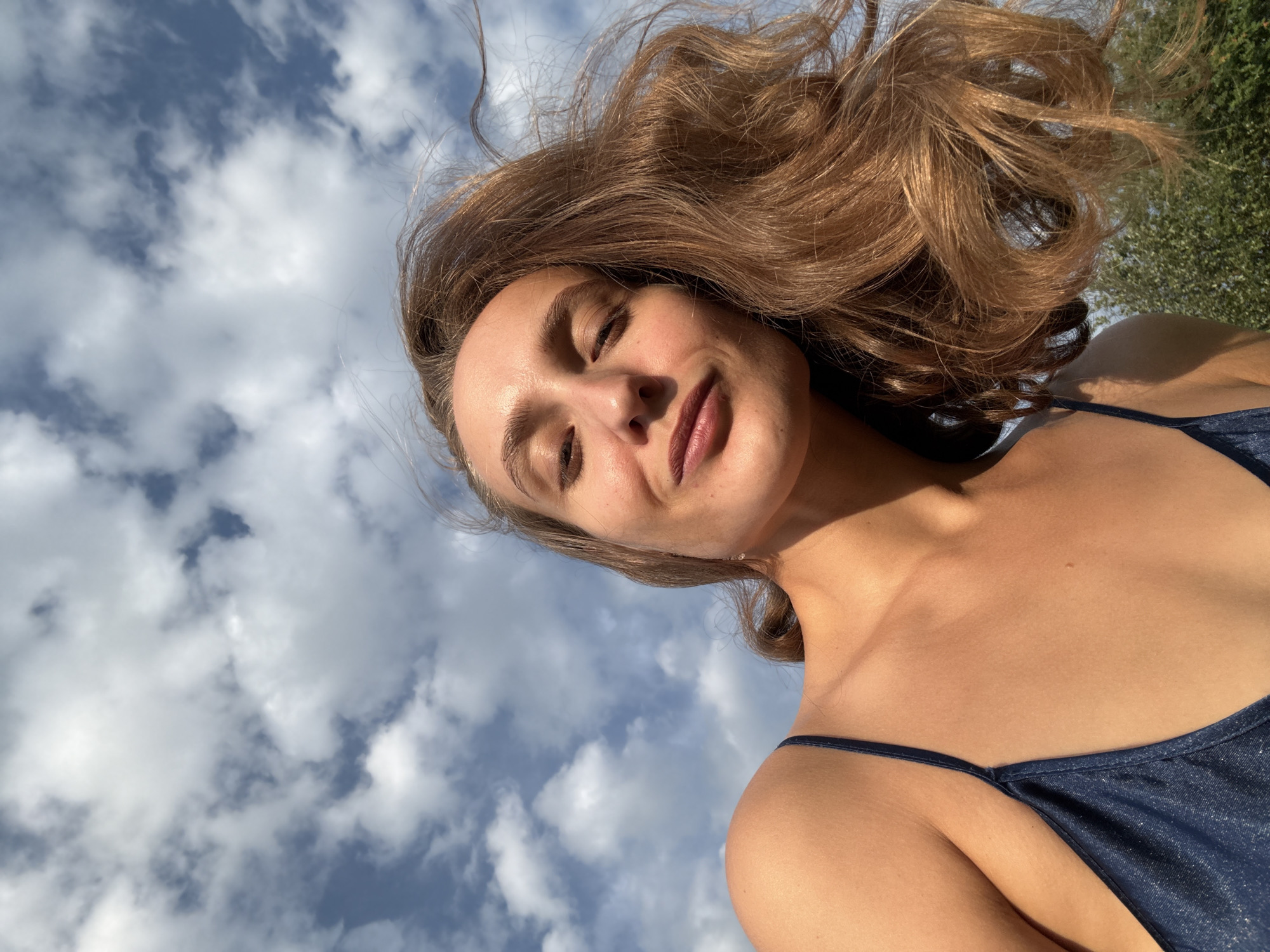 A low-angle, close-up outdoor shot features a woman from the shoulders up, with her face angled towards the camera and eyes mostly closed. She has light brown hair that is windblown, and she is wearing a dark blue, sparkly camisole. The background consists of a bright blue sky filled with fluffy white clouds, with the upper branches of green trees visible in the lower right corner. The lighting suggests it is daytime. There is no information indicating the location in the city of Sharkan, Russia.