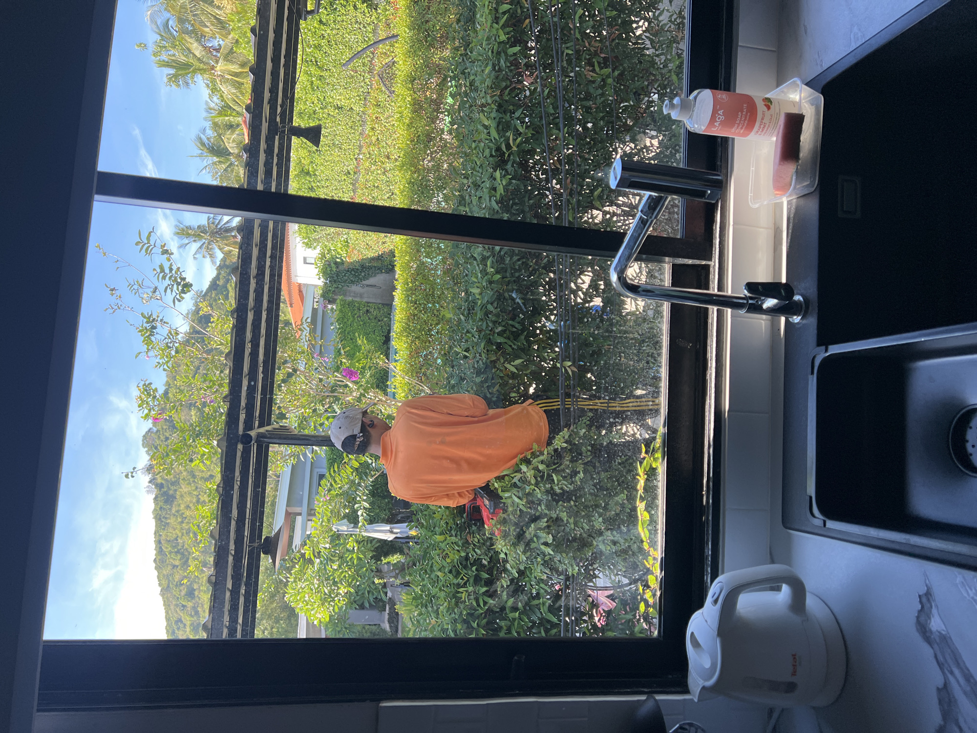 A person wearing an orange shirt and a grey cap is visible outside the window, working on trimming bushes. The window is dark framed and appears to be in a kitchen, with a black sink, a chrome faucet, soap, and a white kettle visible. Outside, the scenery includes trees, grass, and a glimpse of buildings, consistent with a tropical environment. The location is in บ้านบางรักษ์, Thailand.