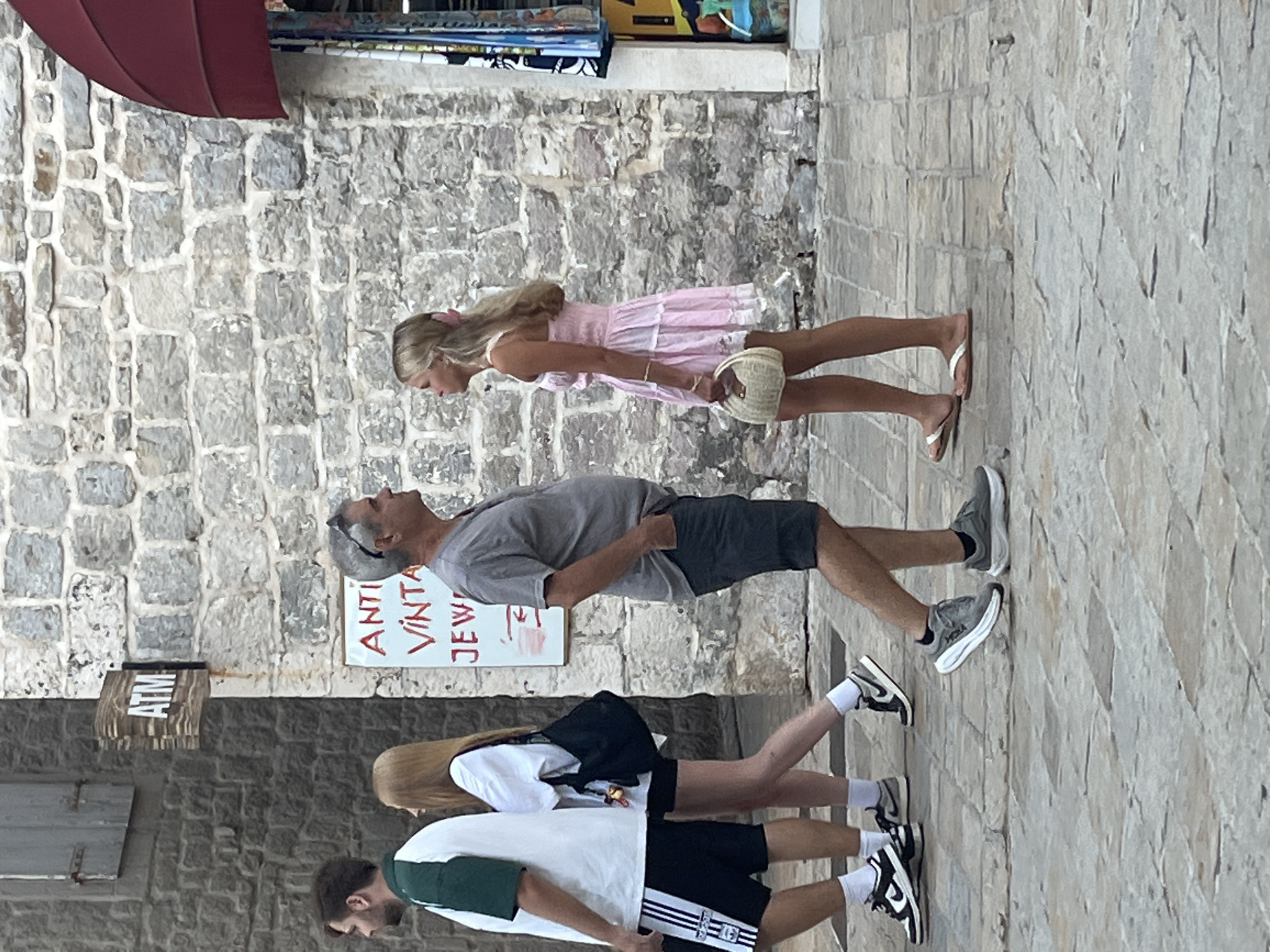 In Budva, Montenegro, a middle-aged man with sunglasses perched on his head and wearing a gray t-shirt and dark shorts stands in profile, looking right. Beside him, a young woman with blonde hair tied with a pink ribbon and wearing a light pink, ruffled dress and flip-flops, also looks right, holding a woven purse. They are walking on a paved stone walkway. Behind them, a man in a green and white shirt and black shorts walks away, and a woman in a white t-shirt and black shorts walks beside him, carrying a black bag. A sign for an ATM is visible on the stone wall to the left, and a sign with red lettering that appears to read "ANTI VINTA JEW" is also on the wall. The architecture consists of stone buildings.