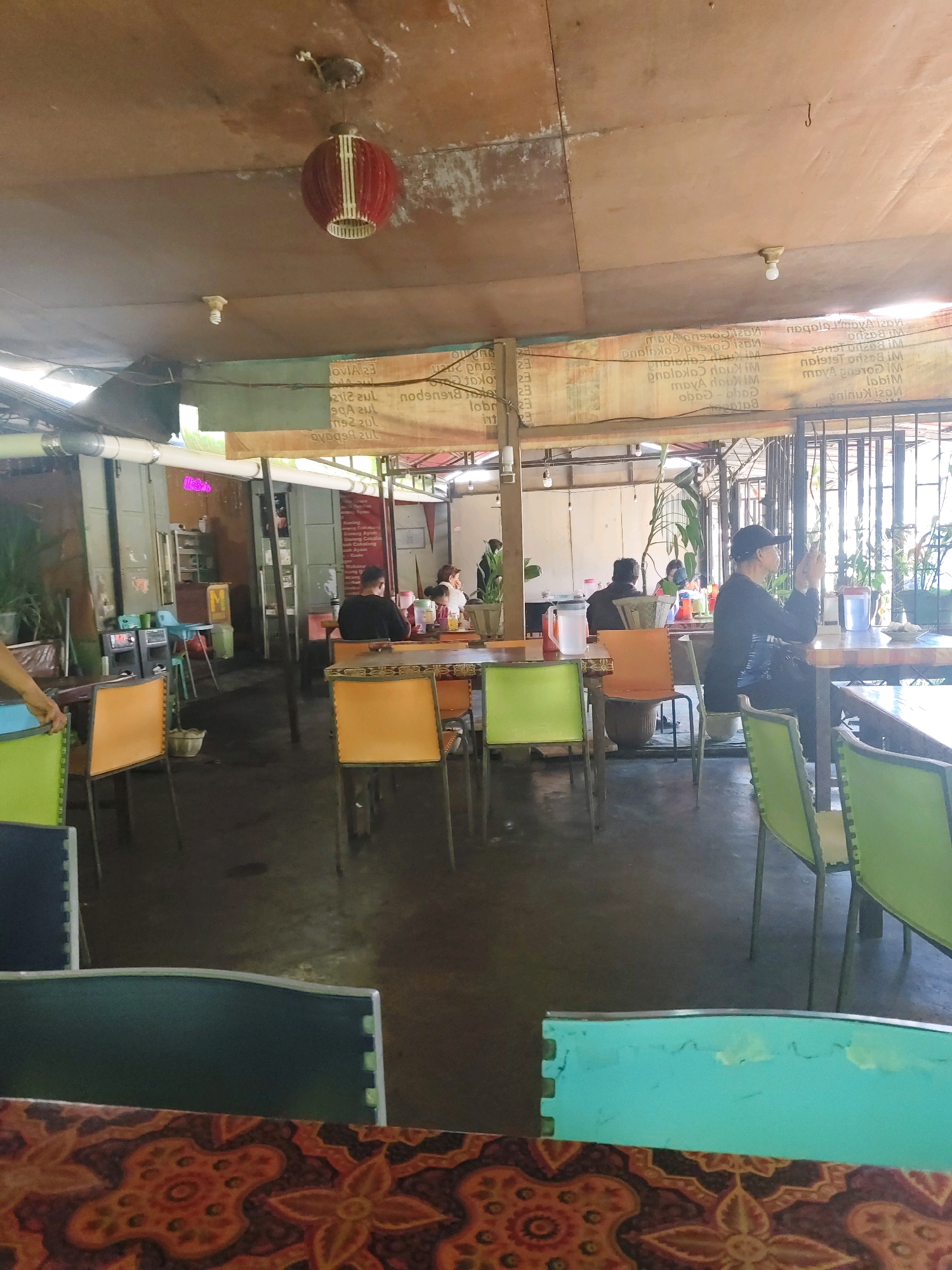 This image depicts the interior of a casual dining establishment, likely a restaurant or eatery, situated in the city of Tomohon, Indonesia. The scene is characterized by a rustic and somewhat unfinished ambiance, with a prominent ceiling made of what appears to be plywood sheets. A red, cylindrical hanging lamp with white vertical slats is suspended from the ceiling, alongside two exposed light bulbs.
The restaurant is furnished with a variety of colorful chairs and tables. Bright orange and lime green chairs surround wooden tables, some of which are covered with patterned tablecloths. The flooring is dark, possibly concrete or asphalt, and shows signs of wear and some dampness. In the background, a metal gate or fence separates the dining area from what seems to be an outdoor space or another section of the building.
Several individuals are visible, engaged in various activities. In the middle ground, a man wearing a black shirt is seated at a table. Further back, a woman in a white top is also seated. To the right, a man wearing a black cap and a black long-sleeved shirt is seated at a table, looking at his phone. He appears to be a patron. Other patrons are visible in the background, some seated at tables, suggesting ongoing dining activities.
Various objects are scattered throughout the scene, including pitchers of liquids on tables, condiment bottles, and what appears to be a basket on the floor. In the background, shelves are visible, possibly holding kitchen supplies or decorative items. The overall impression is that of a busy, informal eating space in Tomohon.