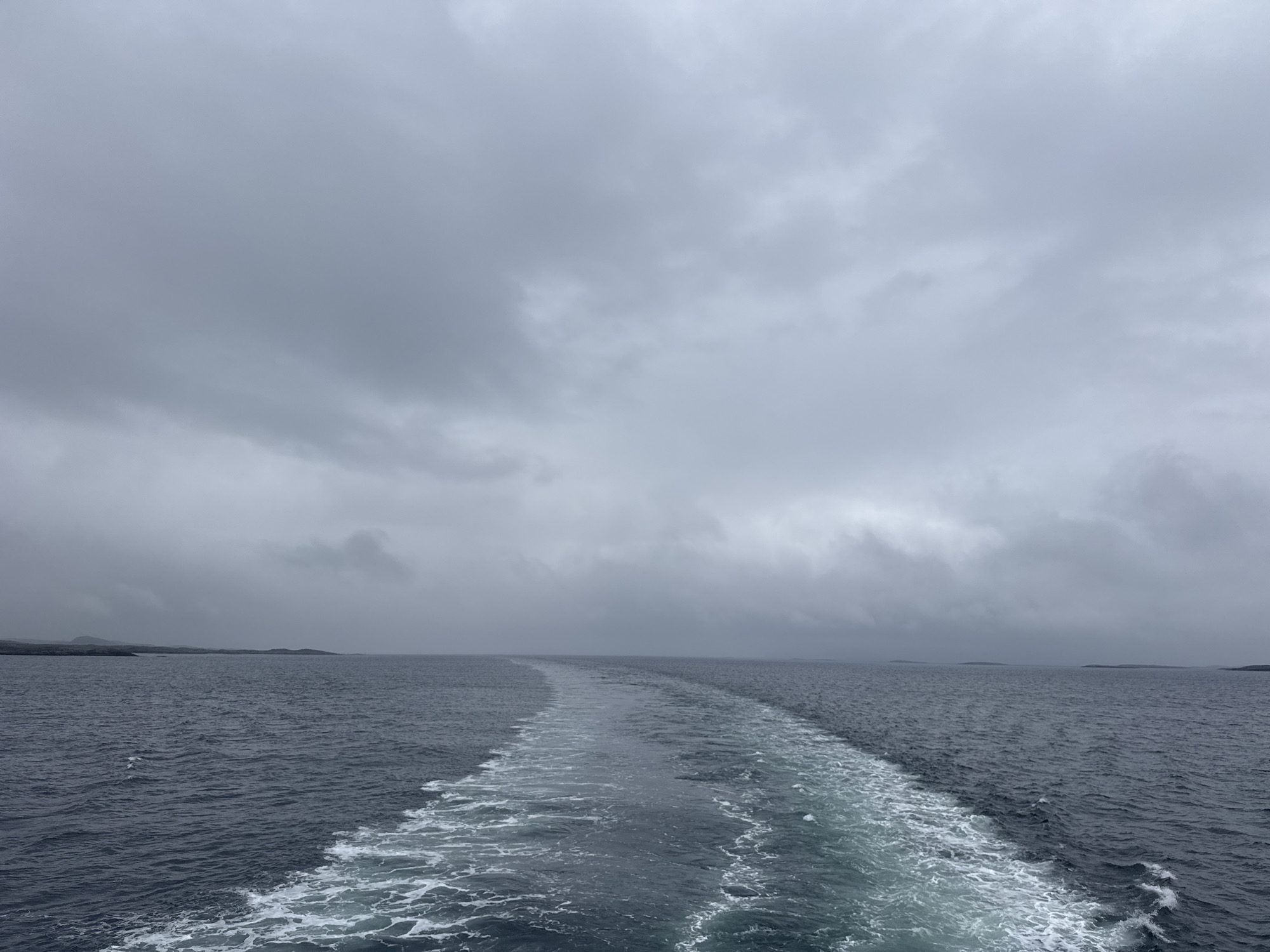 A wide shot captures the open sea under a cloudy sky. The water is a dark, choppy blue. A white wake trails behind the viewer, indicating movement through the water. In the distance, a low, dark landmass is visible, appearing as a hazy silhouette against the horizon. The sky is overcast with dense, grey clouds, suggesting a cool or possibly rainy day. There are no people or specific objects visible in the scene, nor any identifiable landmarks that would suggest the city of Onøya, Norway, without further context. The image depicts a seascape with a focus on the texture of the water and the expansive, muted sky.
