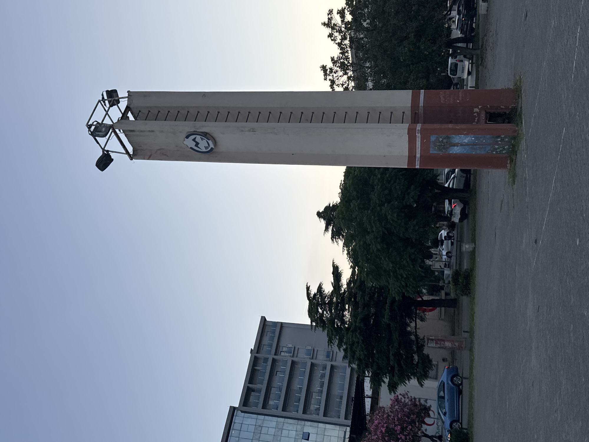 The image depicts a tall, rectangular concrete structure with a light fixture at its top, located in an urban environment in Bar, Montenegro. The structure has a series of metal rods protruding horizontally along its length. A round sign is affixed to its side. The base of the structure has red paint and graffiti-like markings.
The background includes an apartment building with multiple stories, several trees, and parked cars. A blue car is parked near the base of the structure. The sky is visible above, indicating either sunrise or sunset. The ground is paved, appearing to be a parking lot or similar area.