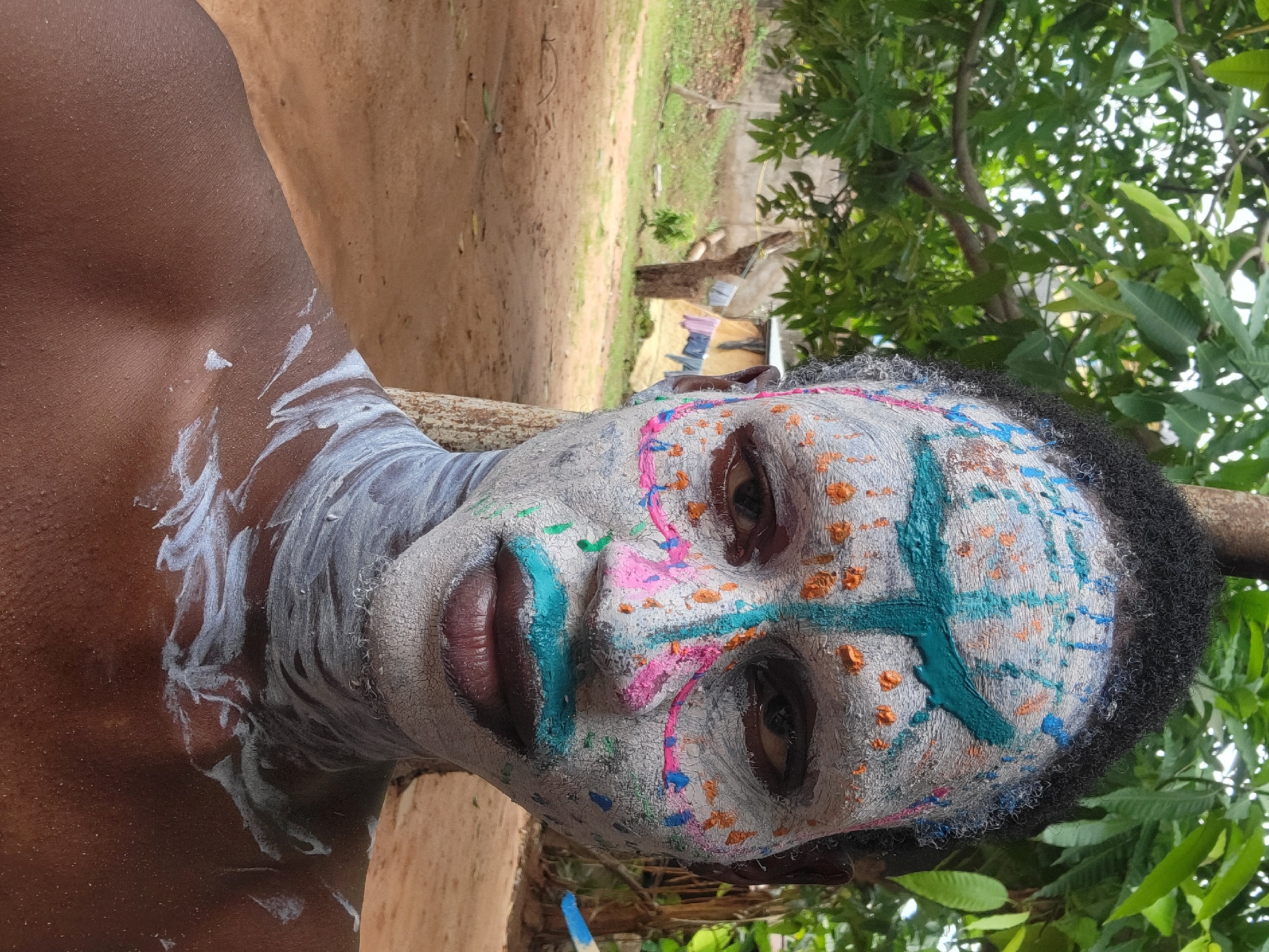 A person with dark skin is close to the camera, their face painted white with colorful markings including blue, pink, green, and orange. The face is partially turned towards the right. The person's lips are painted blue. White paint extends down the neck and onto the upper chest. The background shows an outdoor environment with greenery, dirt ground, and a partial view of a building or structure in the distance. The location is consistent with an outdoor setting in the city of Nkpor, Nigeria.