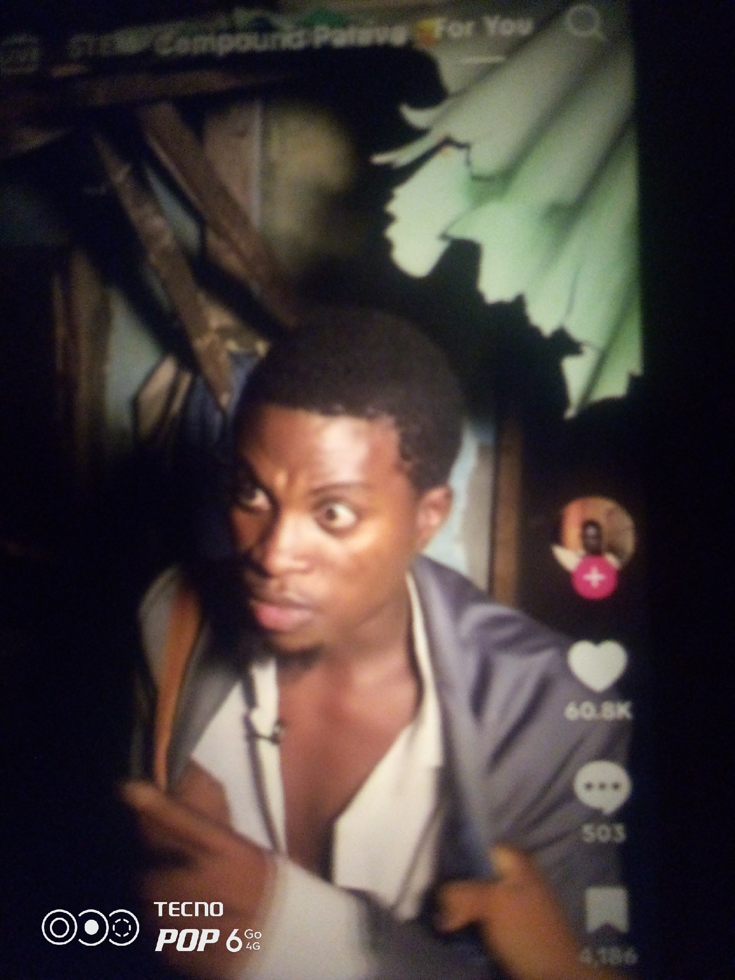 The image shows a close-up of a dark-skinned male with wide eyes, appearing surprised or shocked. He is wearing a grey jacket over a white undershirt. To the right of the subject, there are icons and text overlayed on the image, suggesting it is a screenshot from a social media platform like TikTok or Instagram. The icons include a heart with "60.8K" below it, a speech bubble with "503" below it, and a bookmark icon with "4,186" below it. There is also a small circular profile picture with a pink plus icon in the upper right quadrant of the image. The background is dark and out of focus, with hints of wooden structures and what appears to be a light green fabric hanging. The bottom left corner of the image displays "TECNO POP 6 4G" branding. The image is a photograph taken of a screen, likely a mobile phone. There is no identifiable information within the image that places the scene in Howayi, Nigeria.