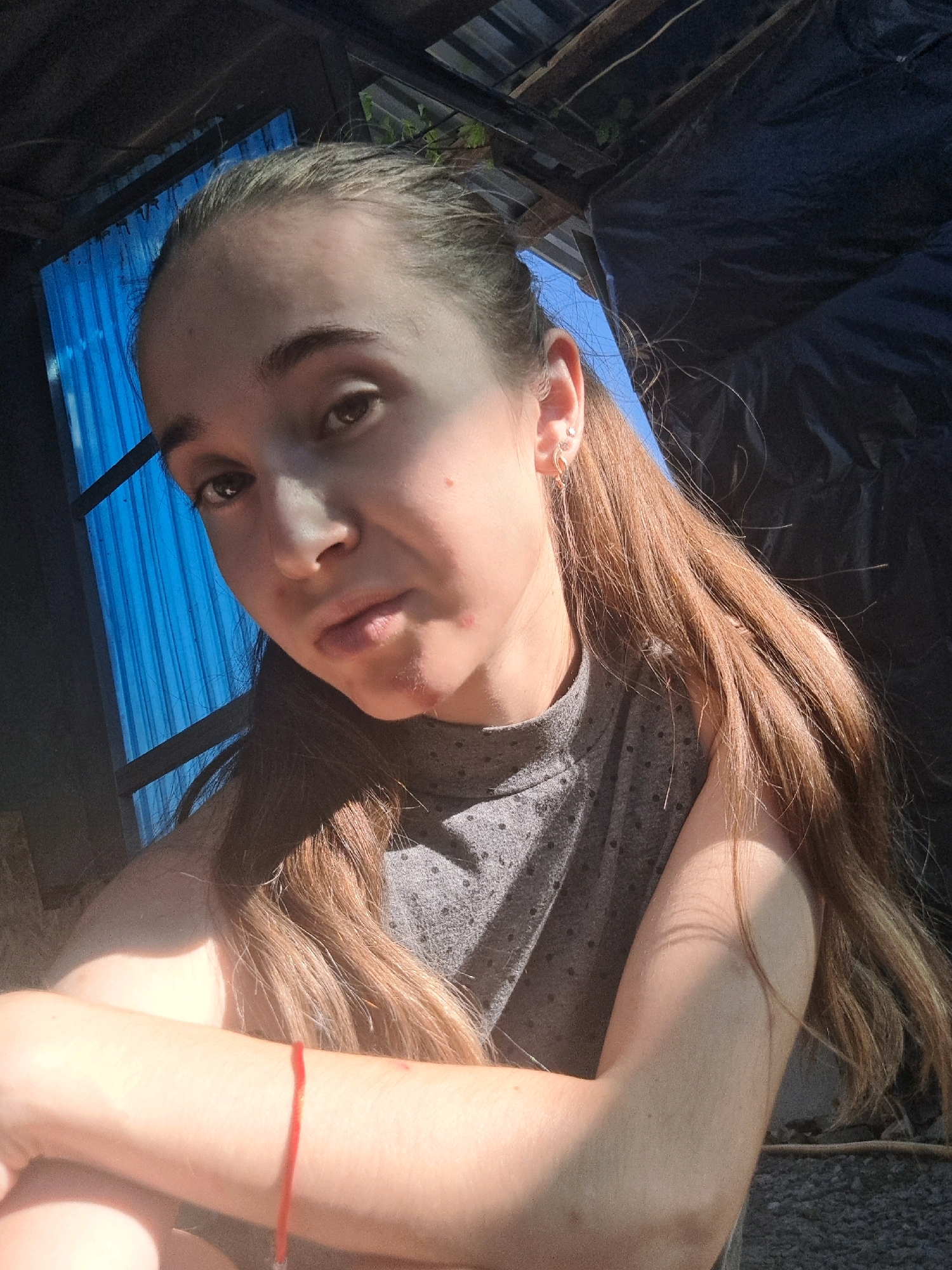 A young woman with long, light brown hair tied into a ponytail is pictured in an outdoor setting, possibly in the city of Vypasne, Ukraine. She is wearing a gray, sleeveless top with a textured pattern. Her arms are crossed in front of her, and she has a red string bracelet on her left wrist. The background is dark and appears to be an improvised structure, with a blue corrugated metal wall on the left side and dark fabric or tarp on the right. Sunlight is illuminating the woman from the side, casting shadows. There are some minor skin blemishes visible on her face.