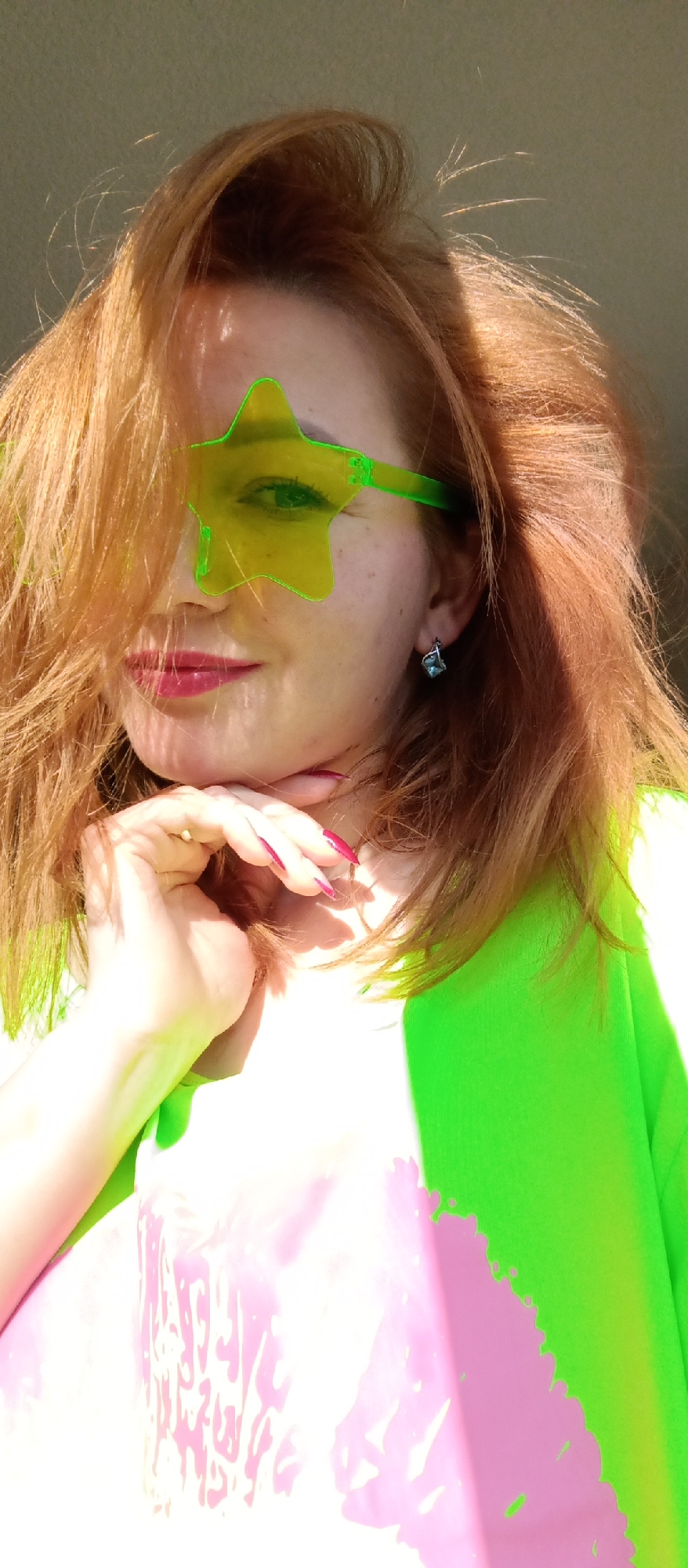 The image shows a person with auburn hair wearing green star-shaped glasses and a bright green top. The person's hand is resting on their chin. The image appears to have a filter or editing applied, resulting in altered colors. The location is unknown.