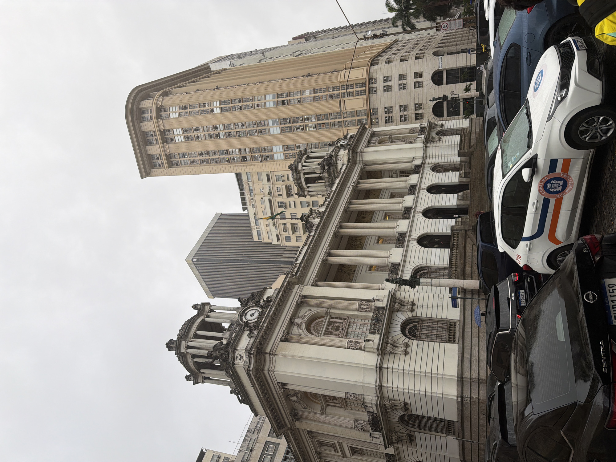 This is a full-frame, eye-level, outdoor, daytime shot of a historic building with classical architecture, likely the Prefeitura (City Hall) of Rio de Janeiro, Brazil, on an overcast day. The building features a dome, columns, and ornate stonework. In the background, several modern high-rise buildings of varying heights and architectural styles are visible. The foreground is occupied by a street with several parked cars, including a white vehicle with official markings that appears to be a municipal service car. One of the cars has a small blue umbrella on its roof. The street is wet, suggesting recent rain. The overall atmosphere is muted due to the cloudy weather.